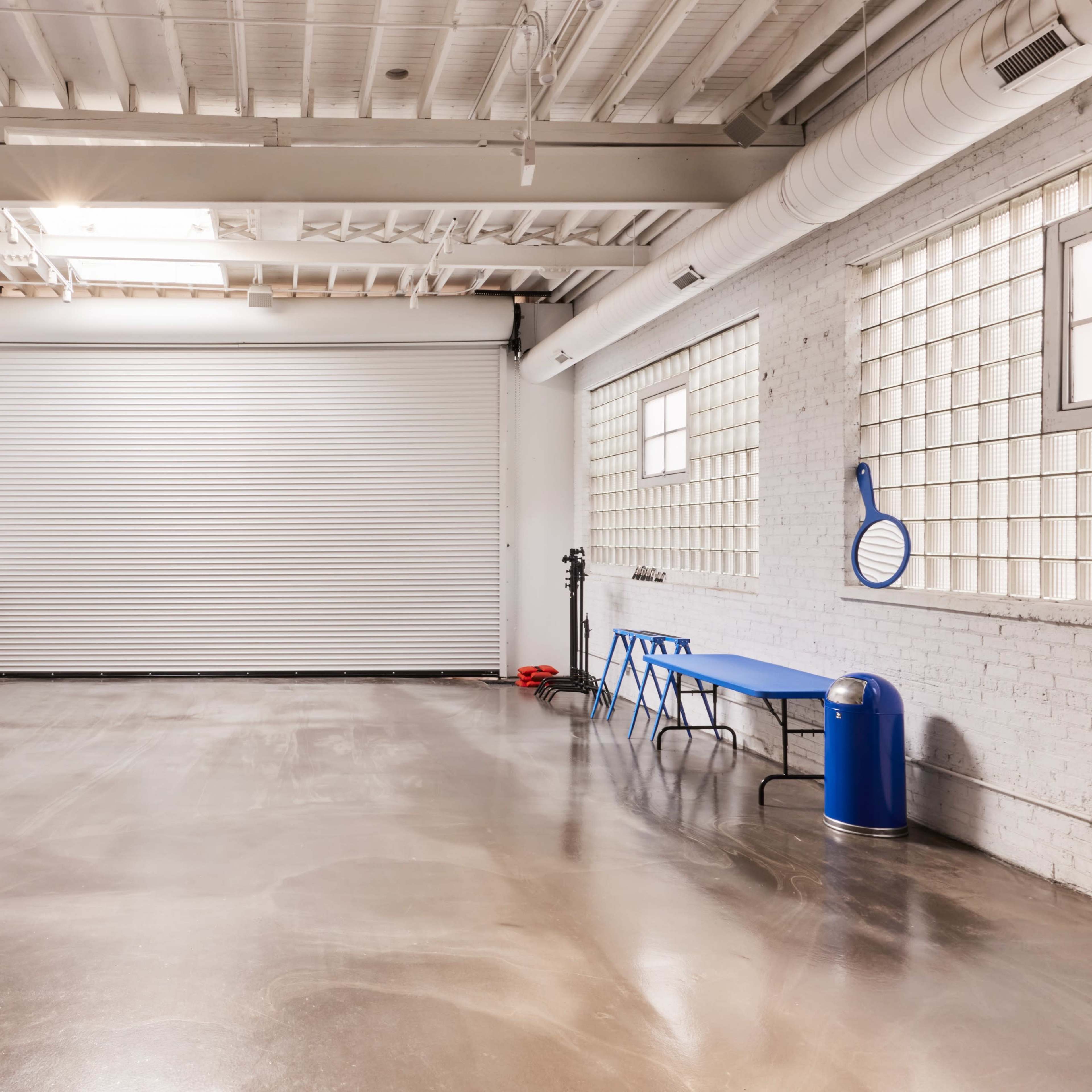 A spacious, minimalistic interior with a roll-up door, a blue table, and a rainbow-colored backdrop stand against a white brick wall.
