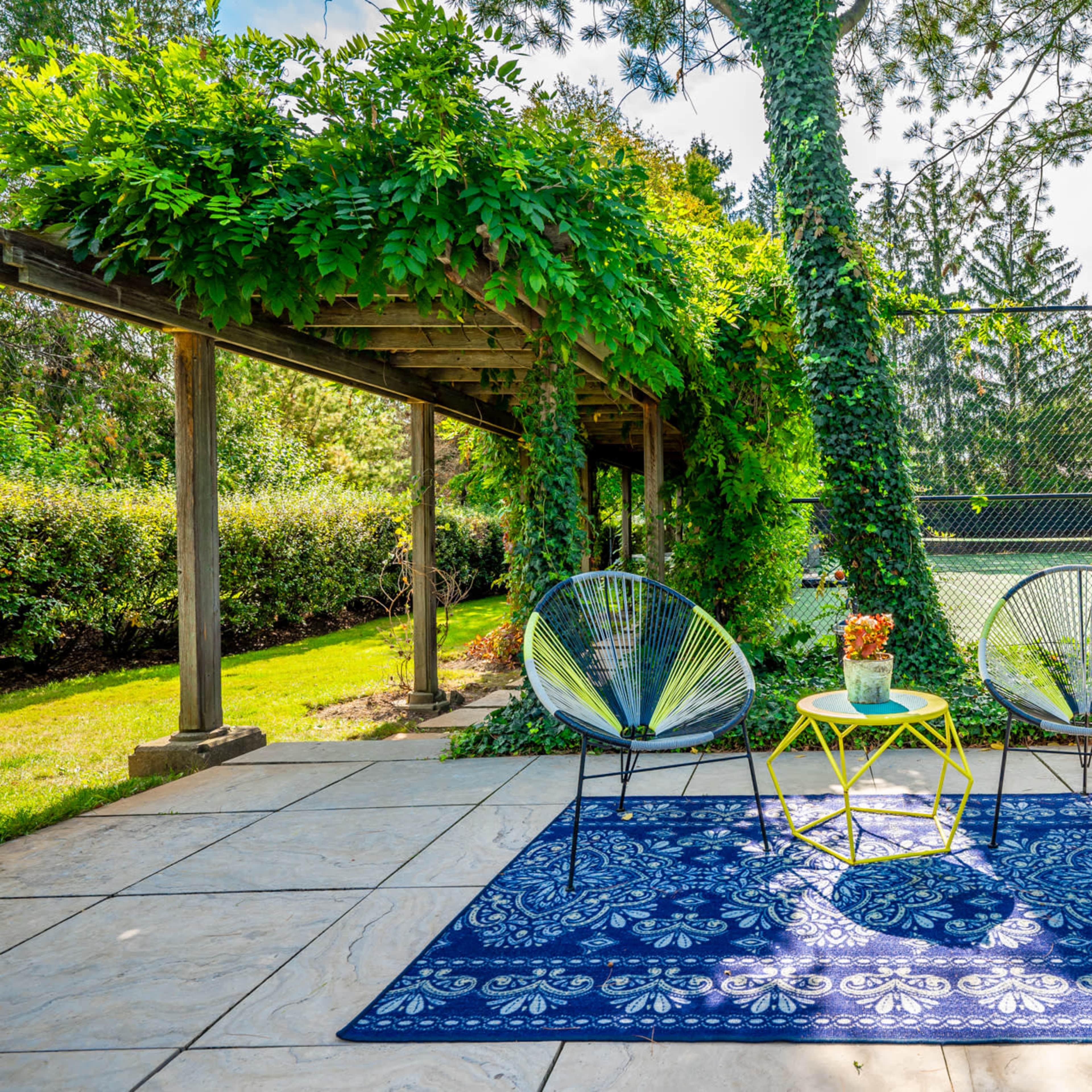 A patio area features two chairs beside a small table on a patterned rug, surrounded by greenery and facing a tennis court in the background.