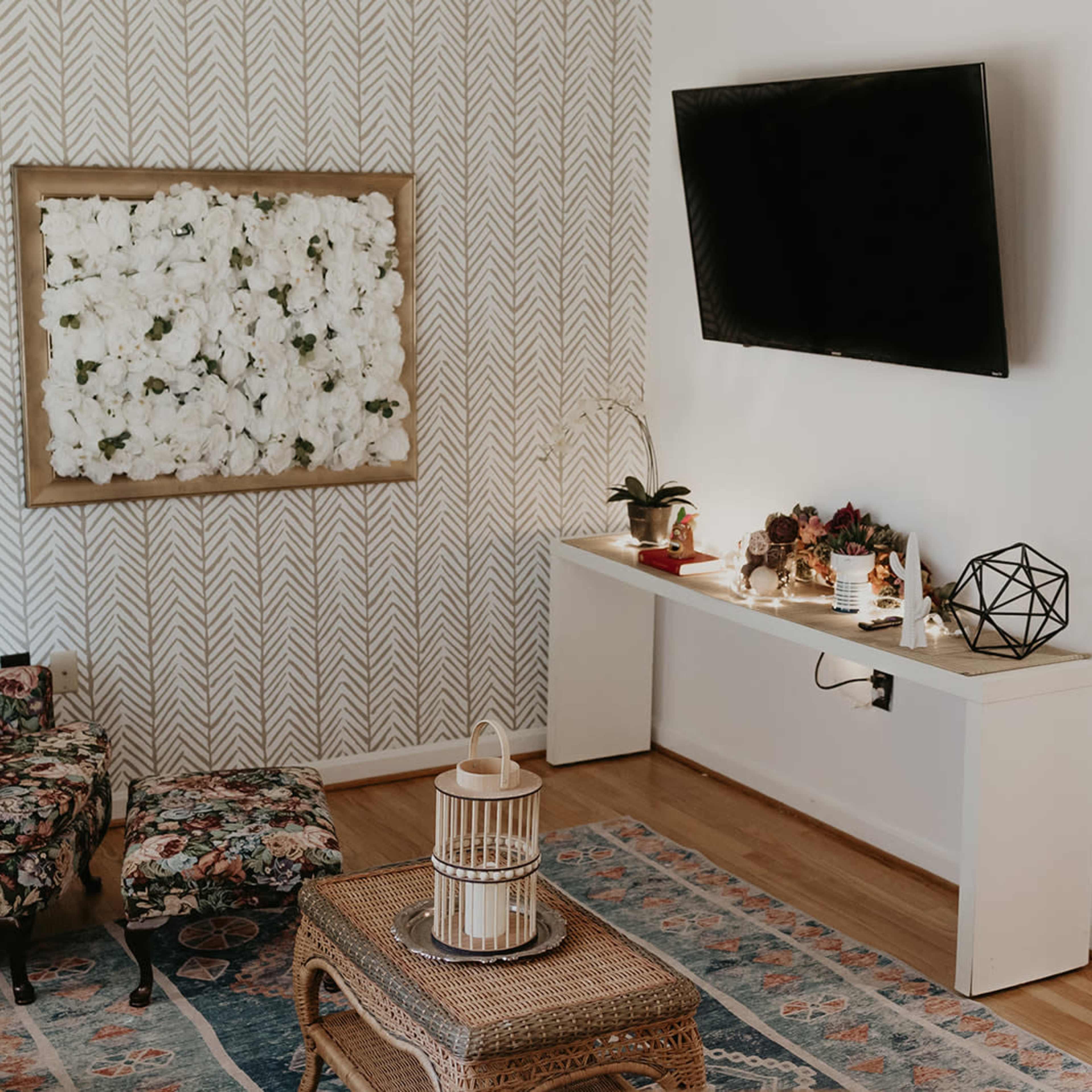 A cozy living room with floral-patterned furniture, a wall-mounted TV, and a console table decorated with plants and decorative objects.