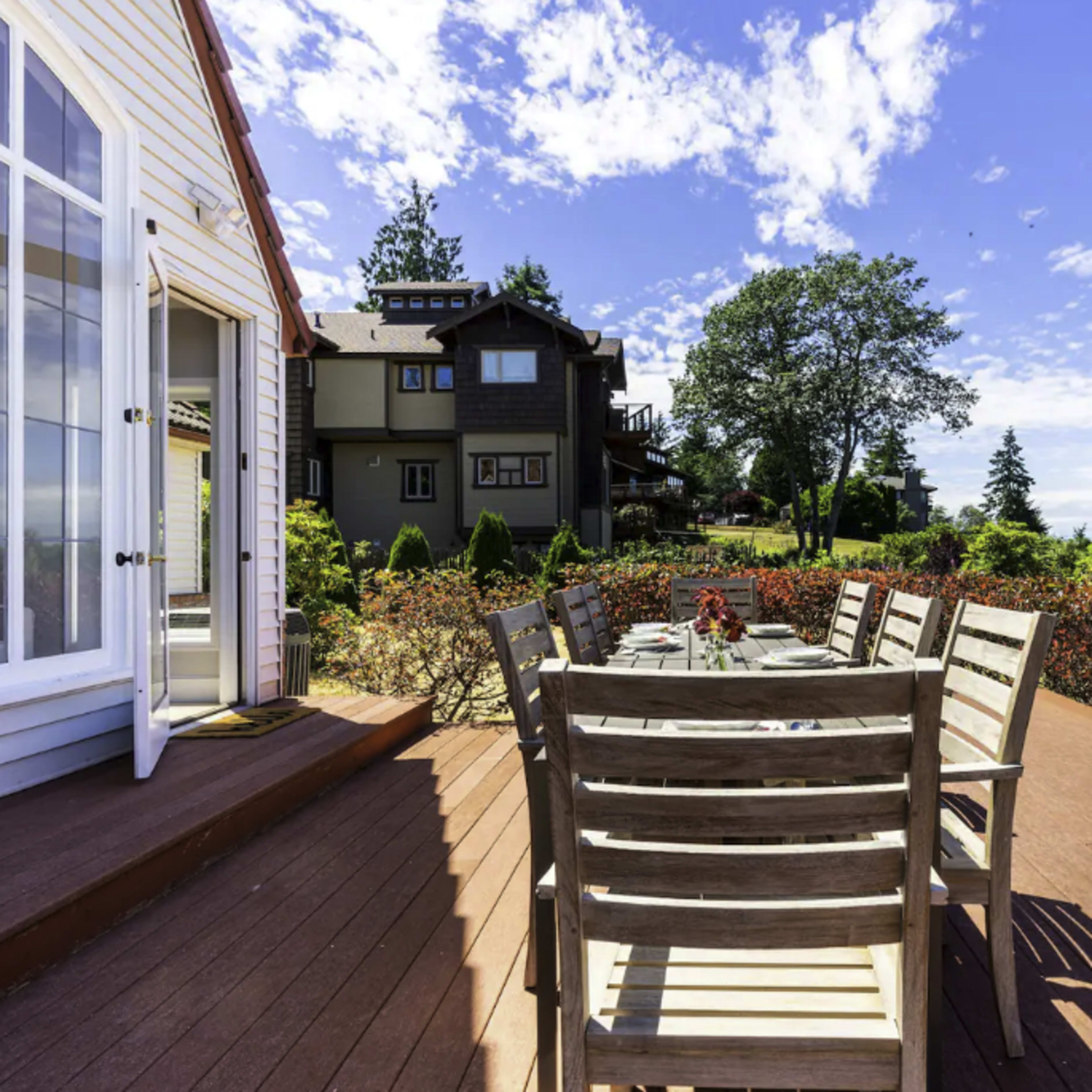 A wooden deck with a large dining table and chairs overlooks a landscaped garden and houses under a clear blue sky.