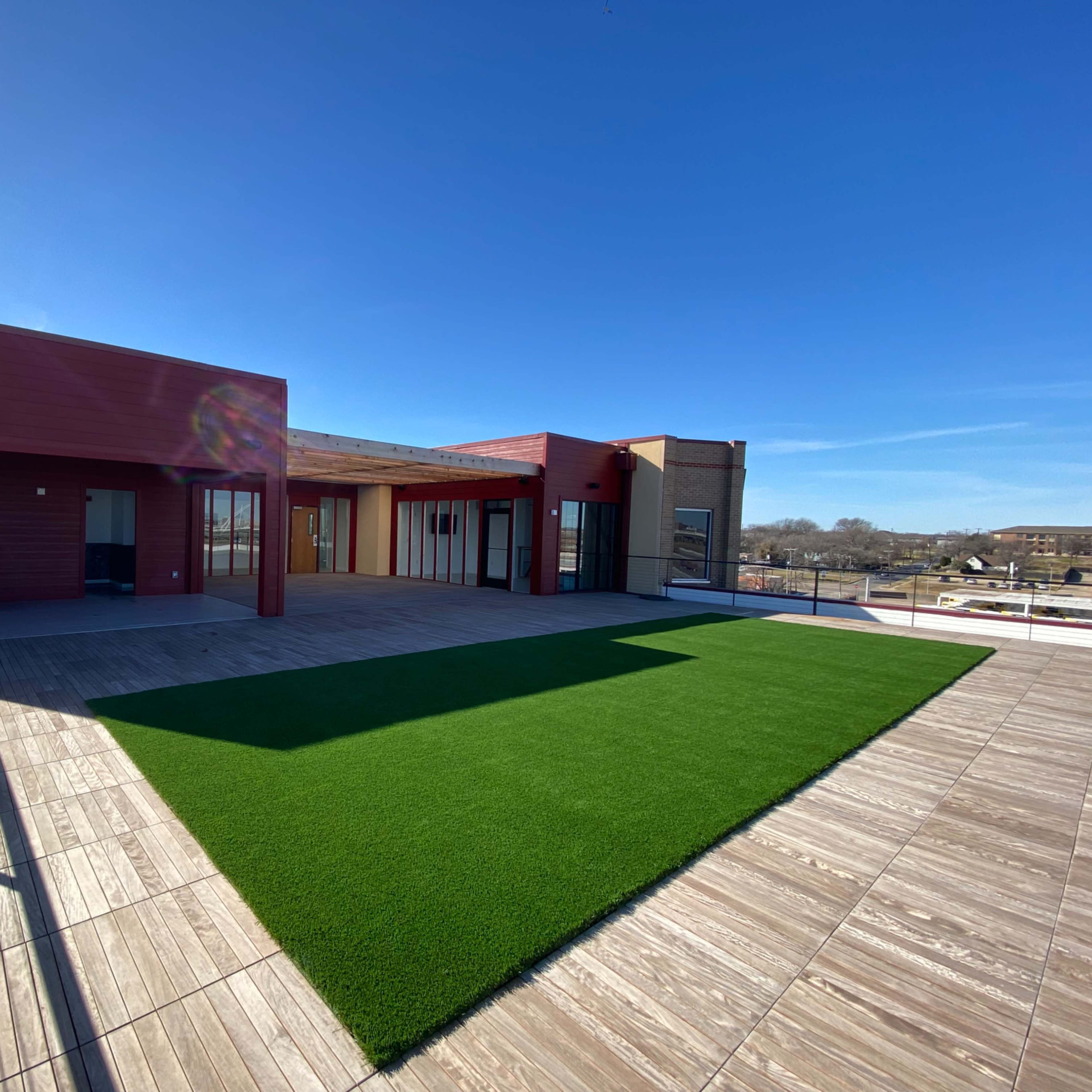 A rooftop space with a green artificial grass area and wooden decking, alongside modern buildings under a clear blue sky.