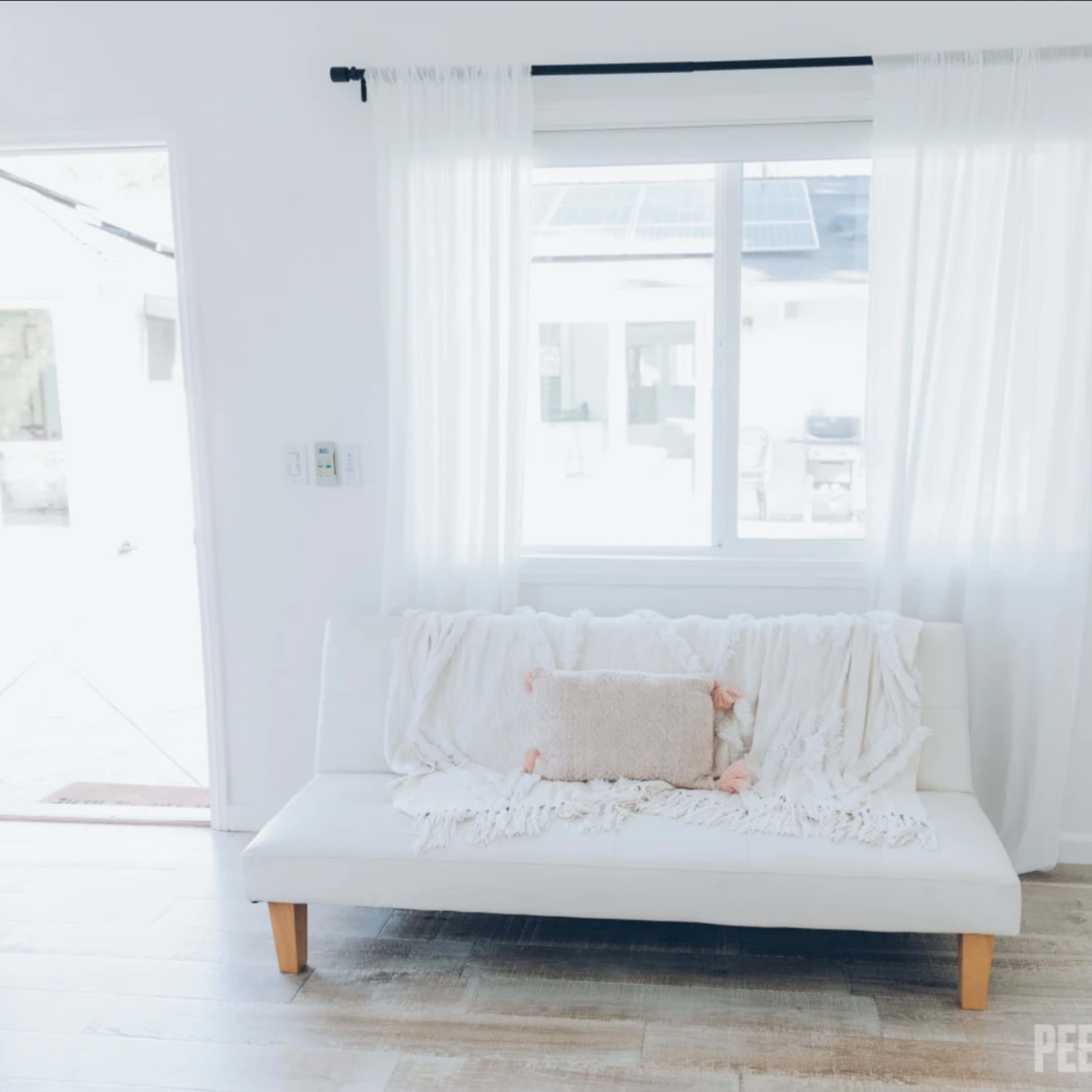A white couch with a light blanket and decorative pillow is positioned in front of a large window with sheer curtains, illuminating a bright, minimalist room.