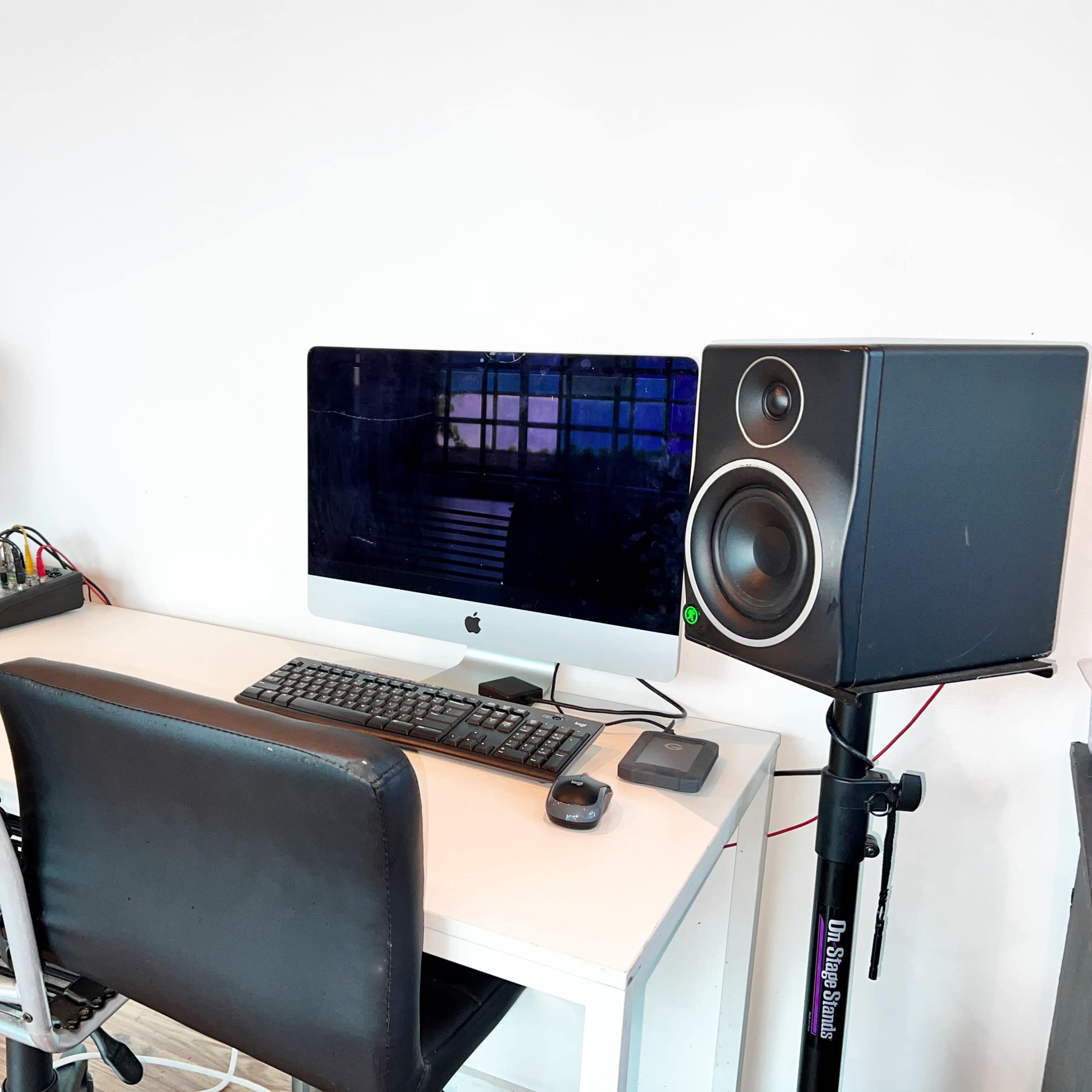 A desk with an iMac, a keyboard, a mouse, and two studio speakers is set up against a white wall.