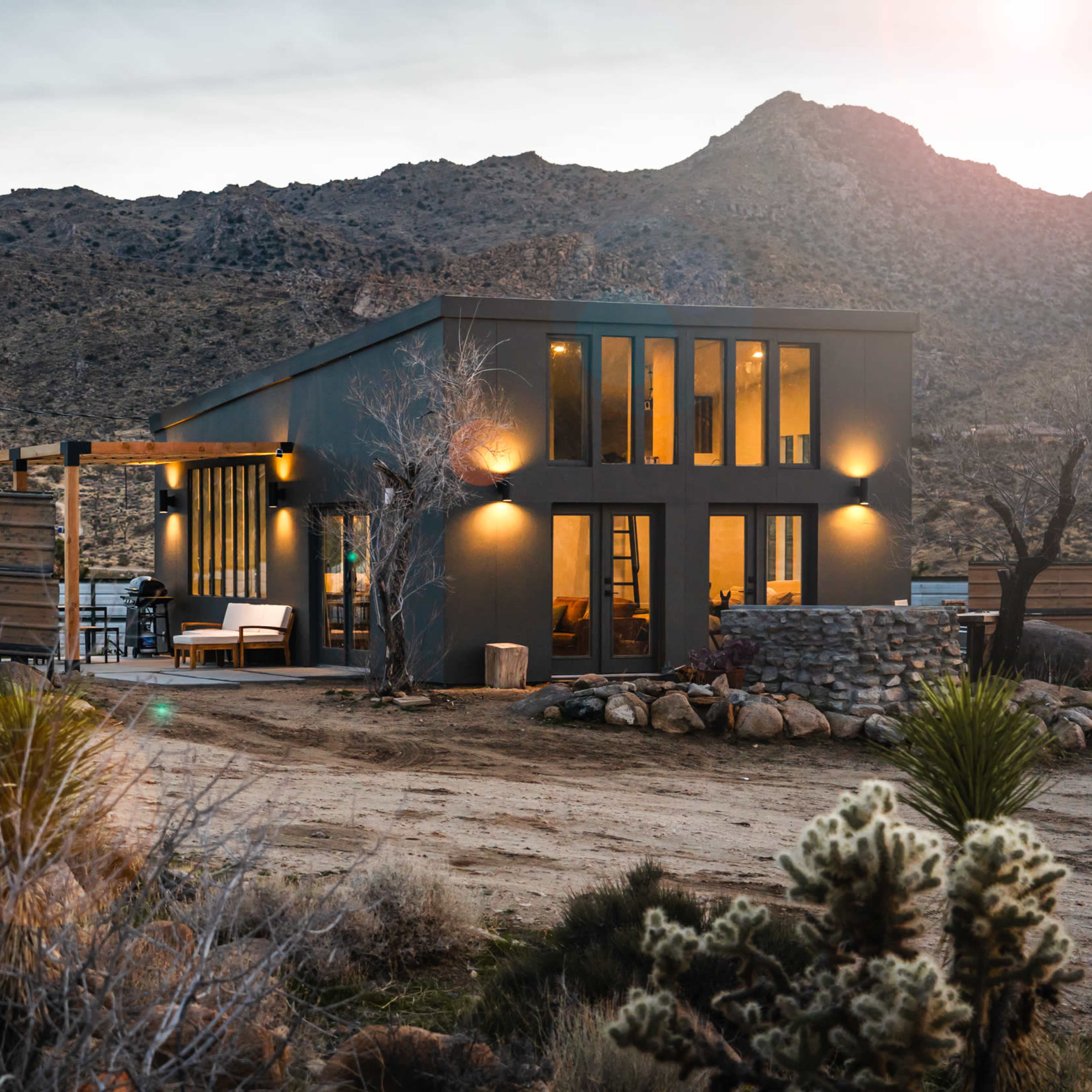 A modern house with large windows is situated in a desert landscape surrounded by rocky hills and sparse vegetation.