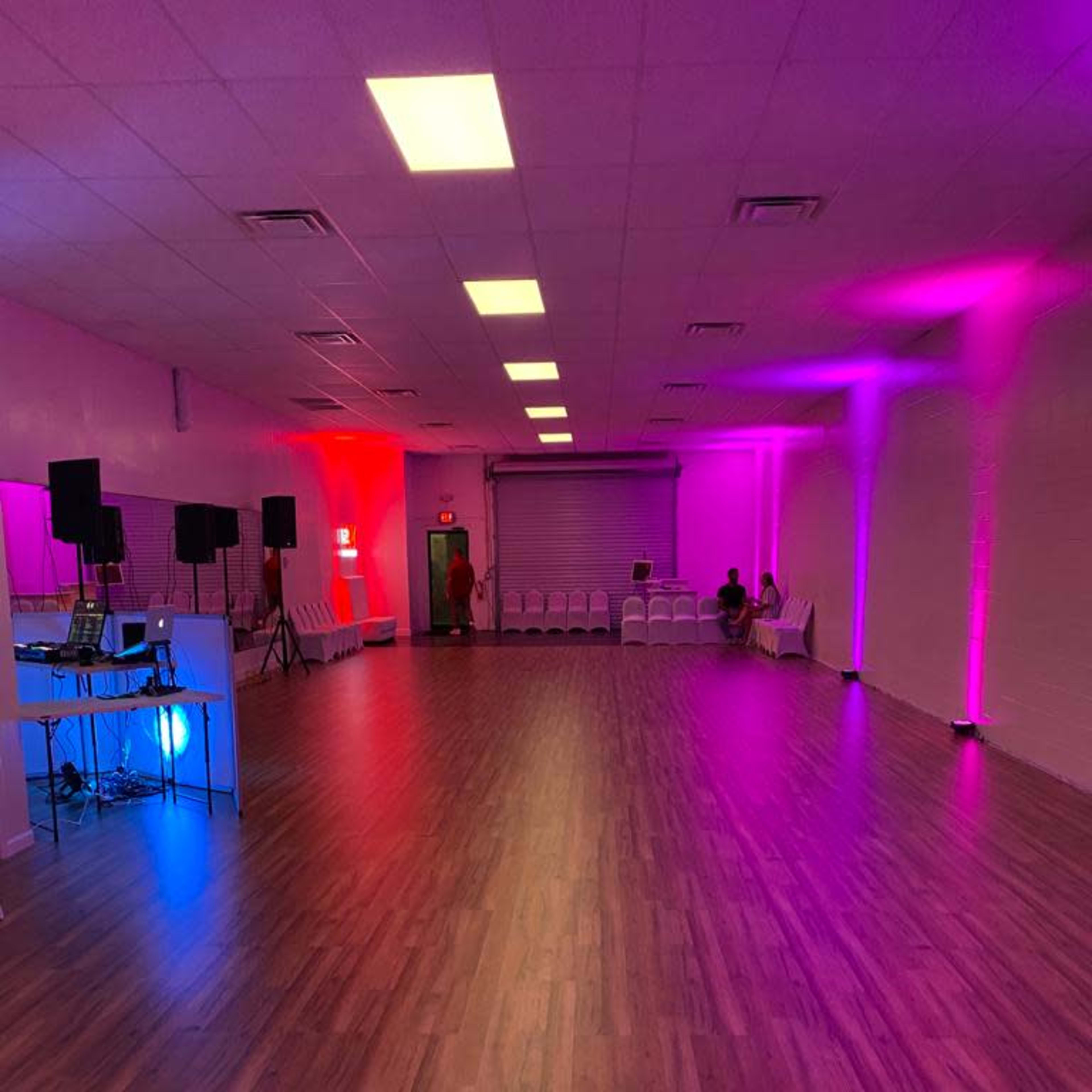 A spacious room with a polished wooden floor, featuring colorful lighting along the walls and a setup for music equipment at one end.