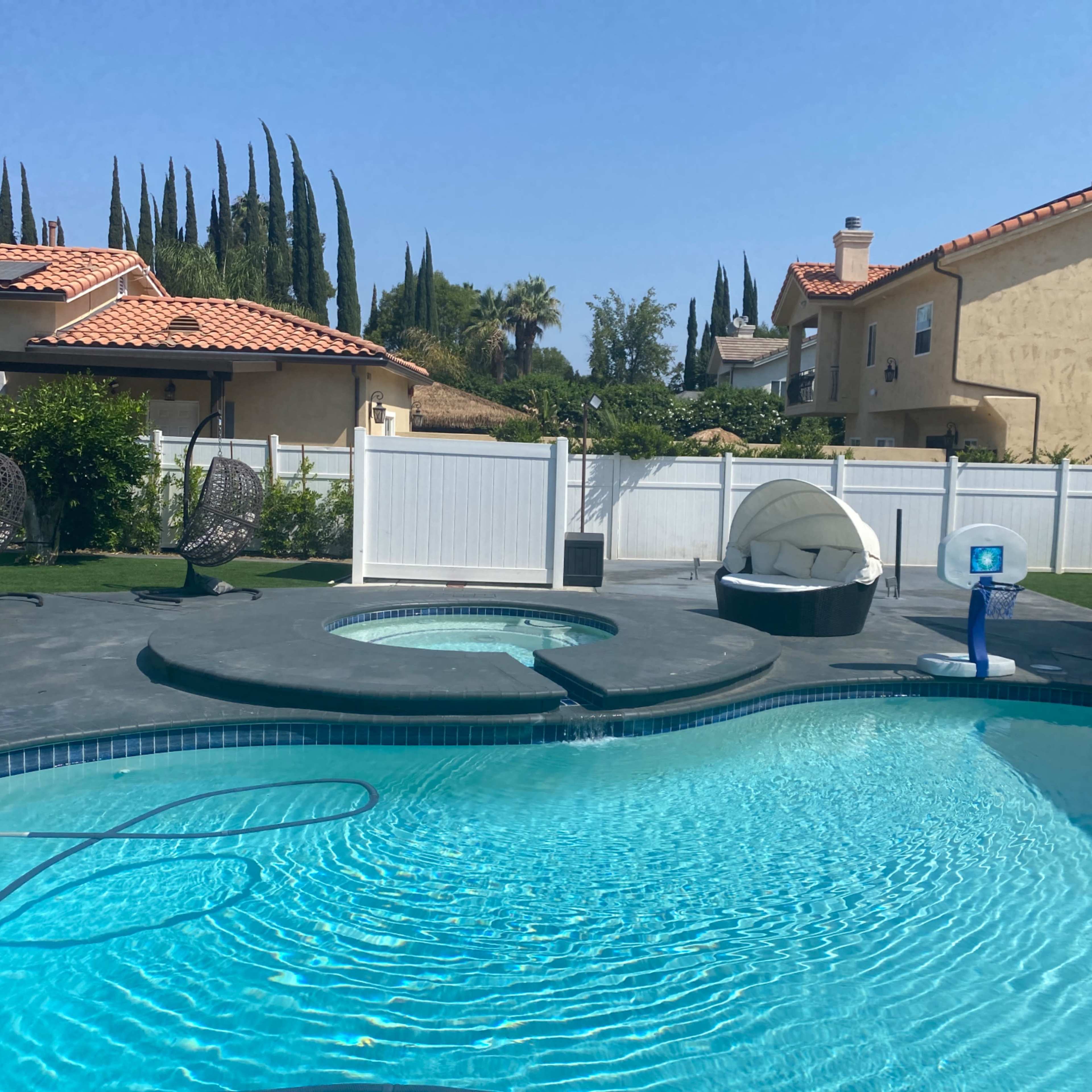 A clear blue swimming pool with a spa, surrounded by a neatly manicured yard and several tall trees in the background.