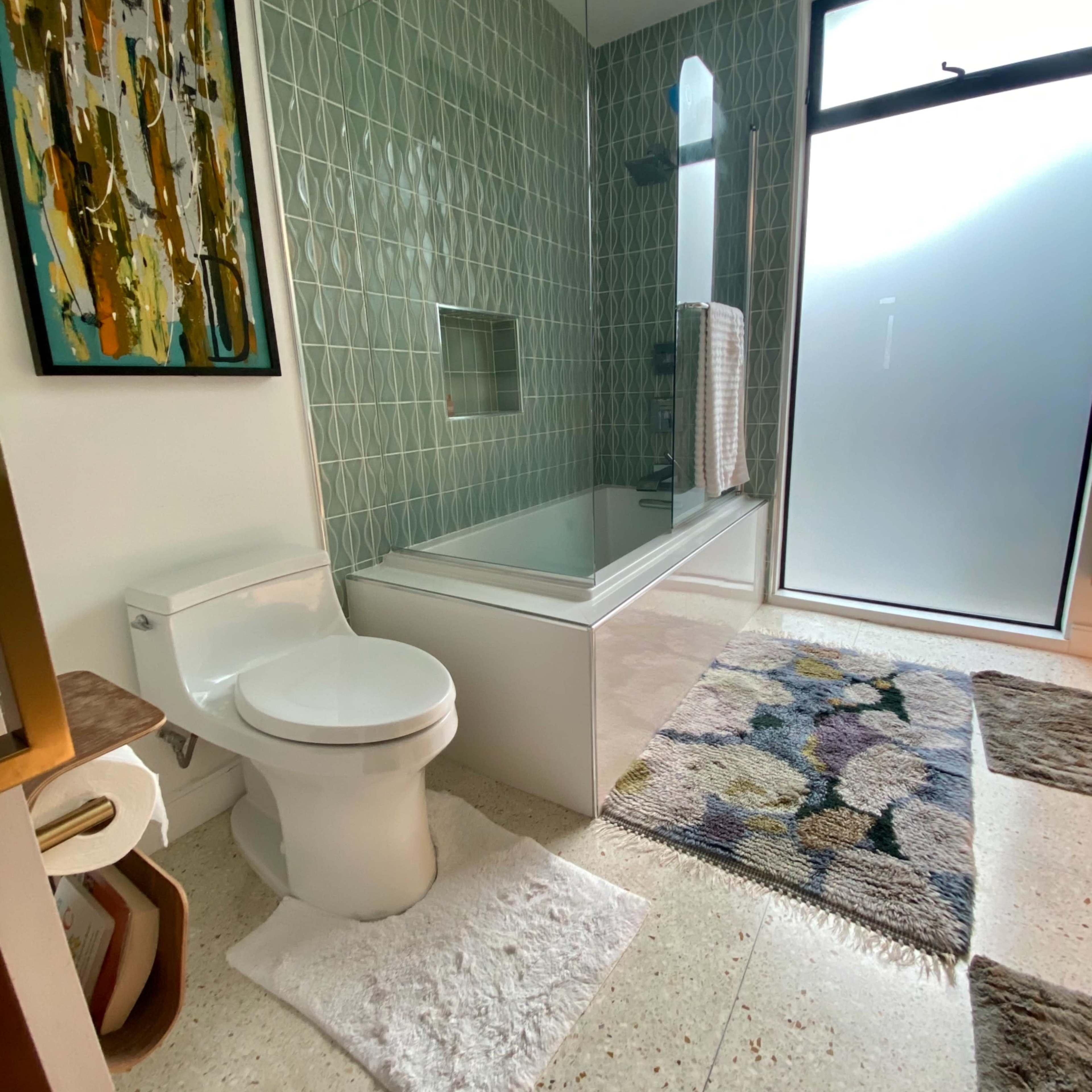 A modern bathroom featuring a toilet, a bathtub with a shower, a patterned rug, and a large frosted glass window.