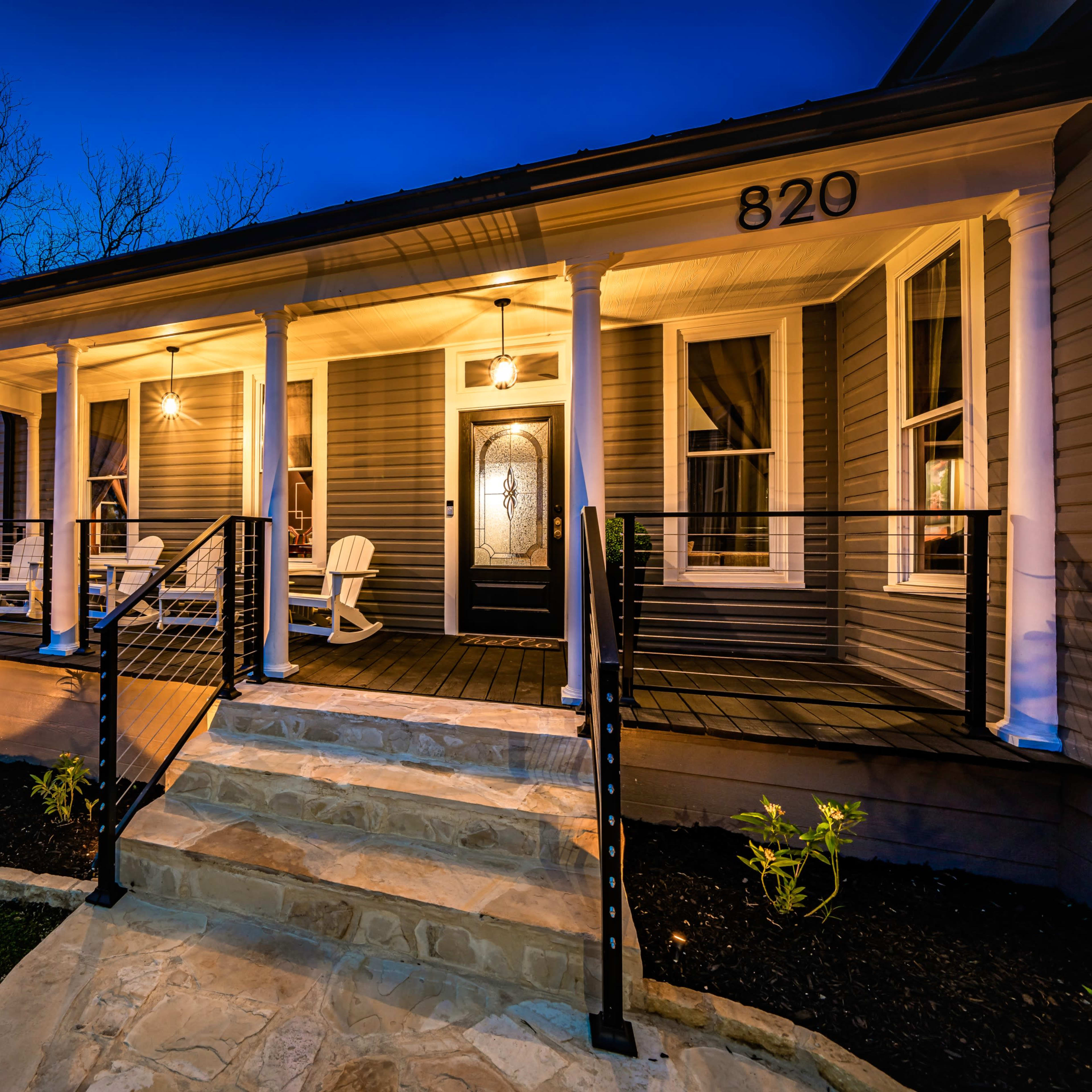 A well-lit porch with white rocking chairs and a stone walkway leading up to a house at dusk.