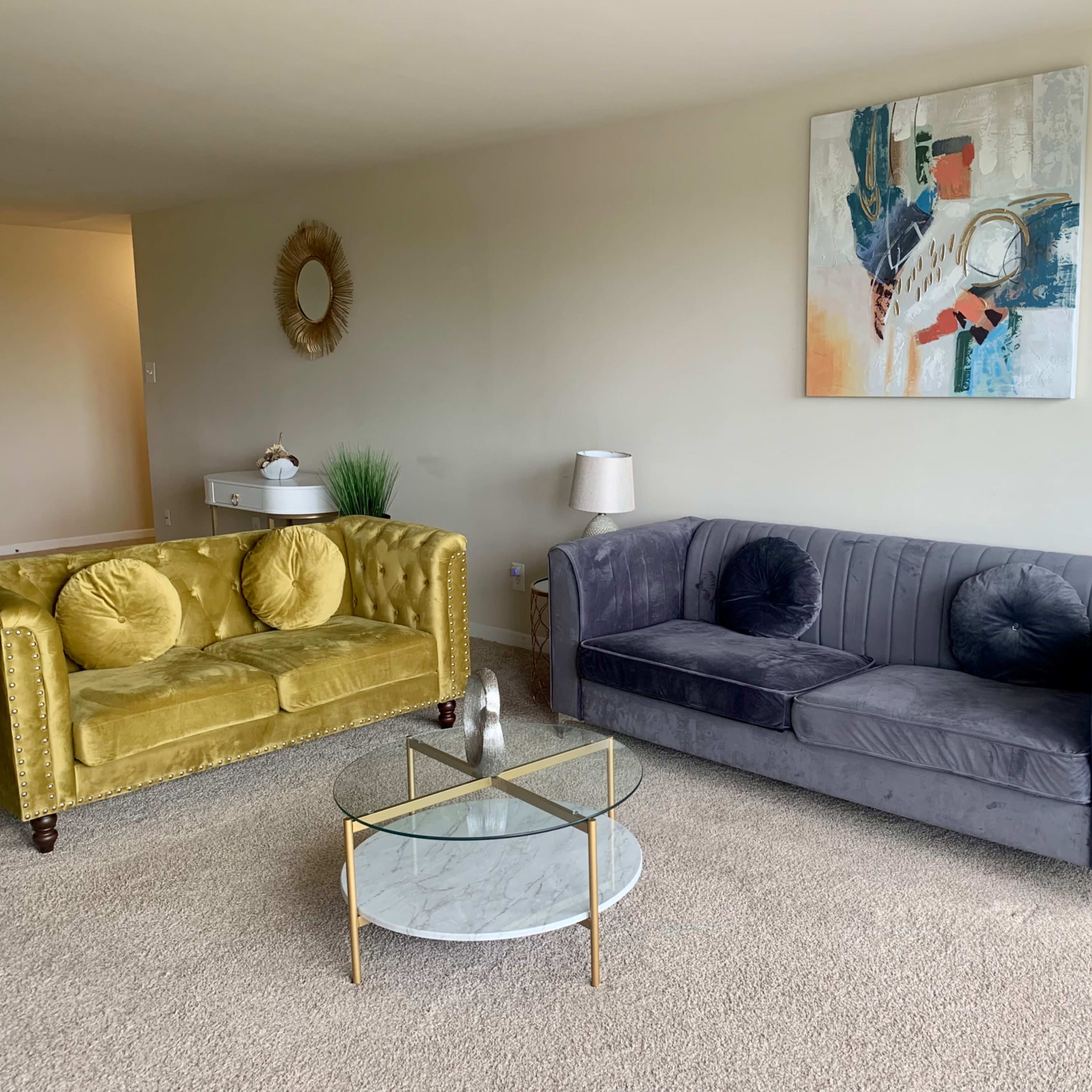 A living room with a yellow velvet couch, a gray upholstered couch, a glass coffee table, and a decorative wall art piece.