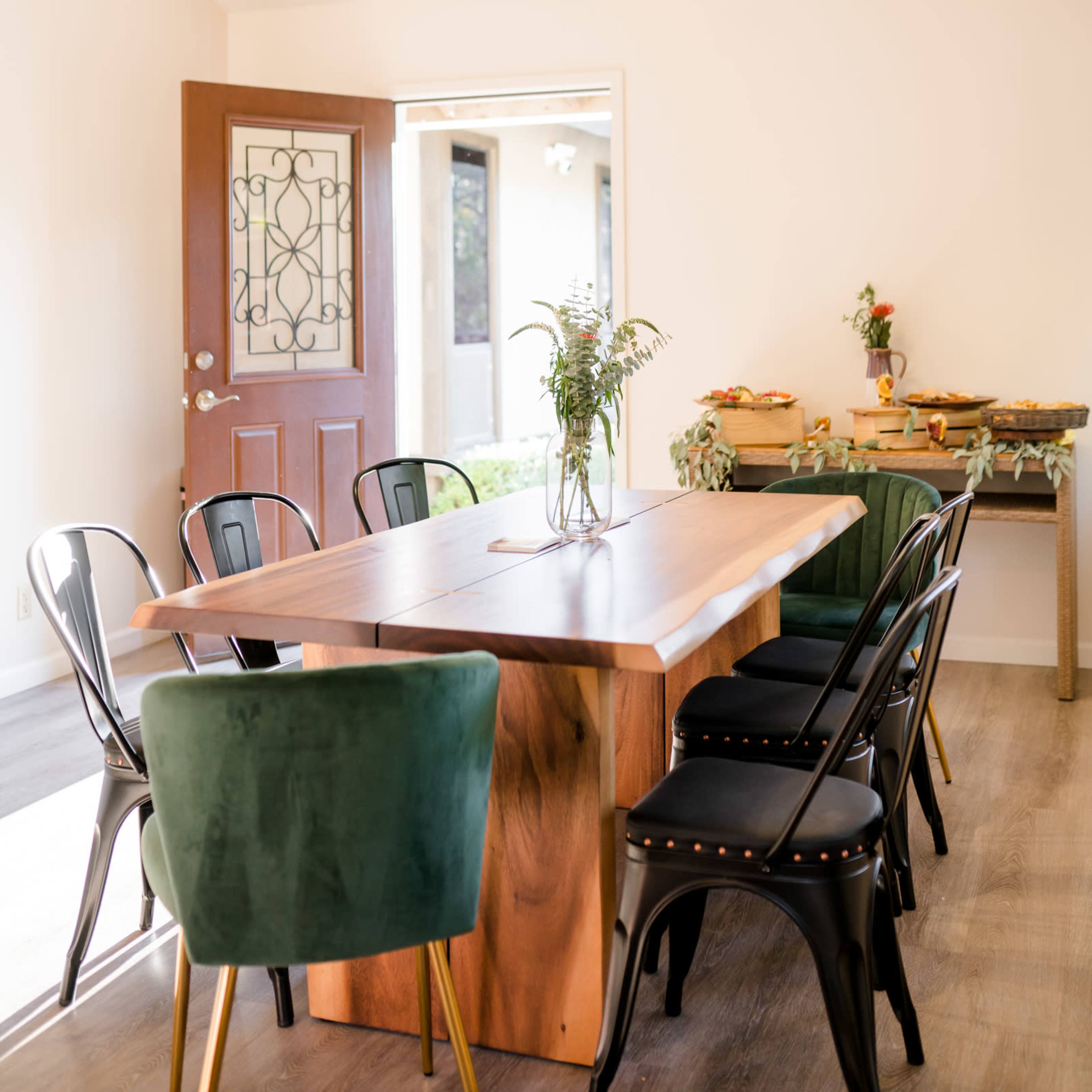 A dining area features a wooden table surrounded by green and black chairs, with a door leading outside and a side table with food.