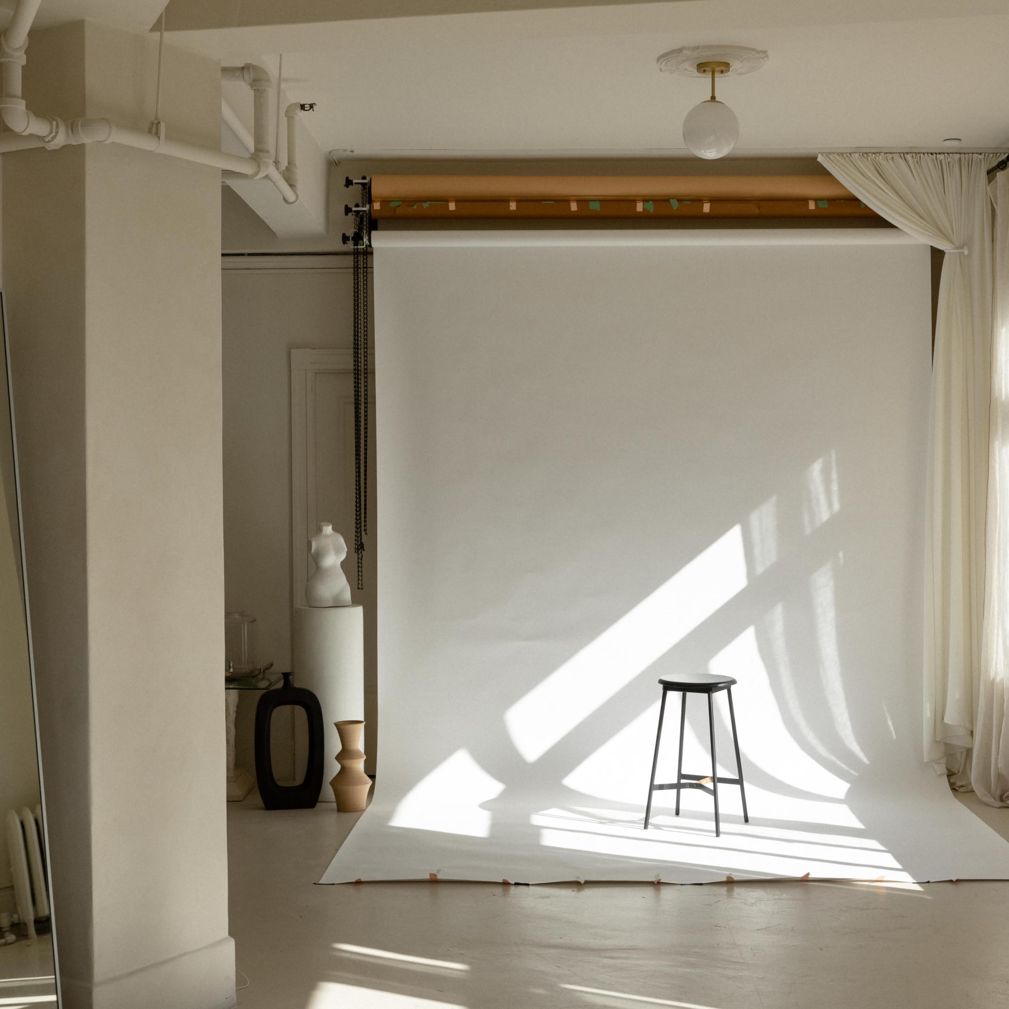 A minimalist studio setting features a black stool positioned on a white backdrop with sunlight casting shadows across the floor.