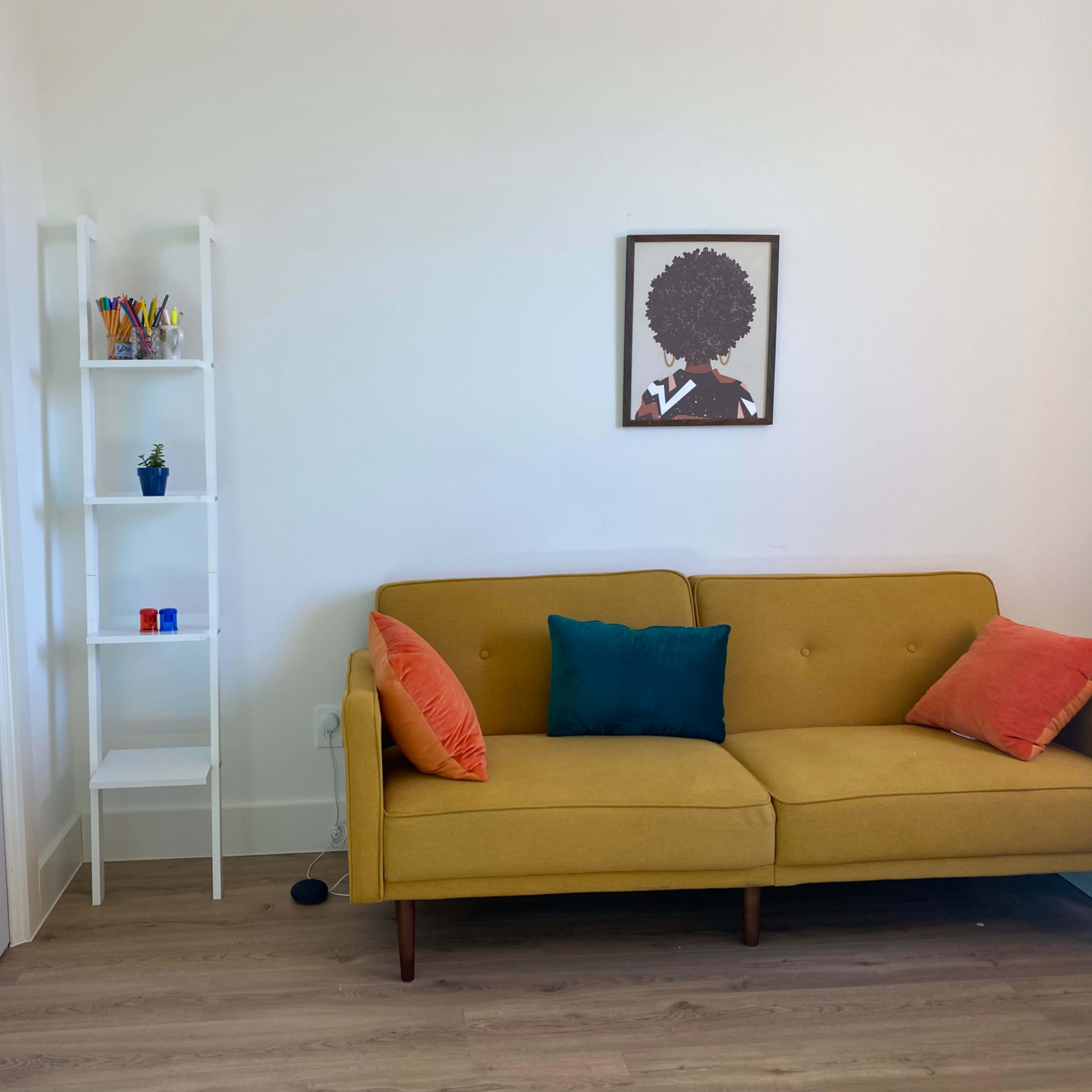 A yellow couch with orange and blue pillows sits against a white wall, next to a white shelving unit with various items and a framed artwork above it.