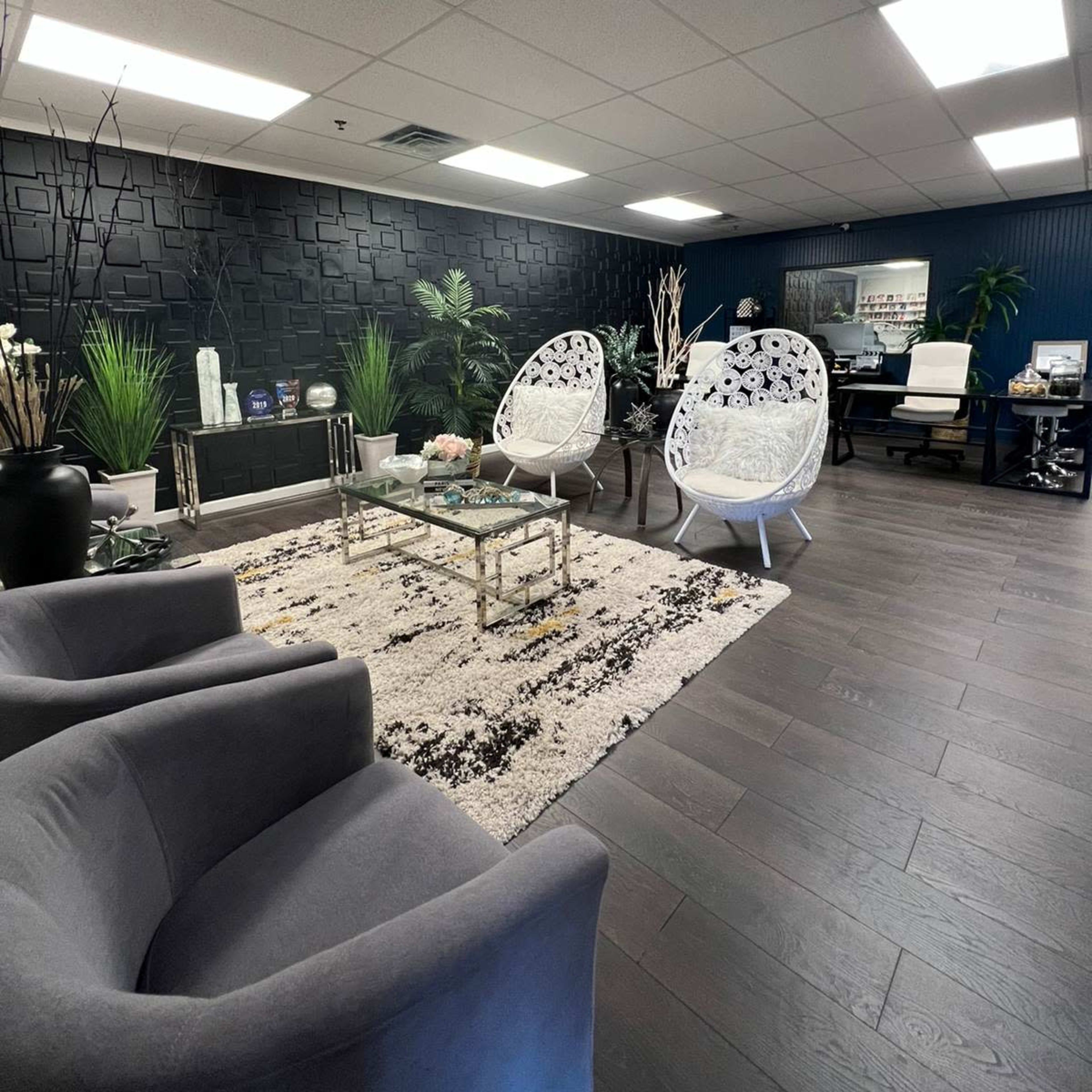 A modern office waiting area with two white chairs, a gray sofa, a glass coffee table, and decorative plants against a dark wall.