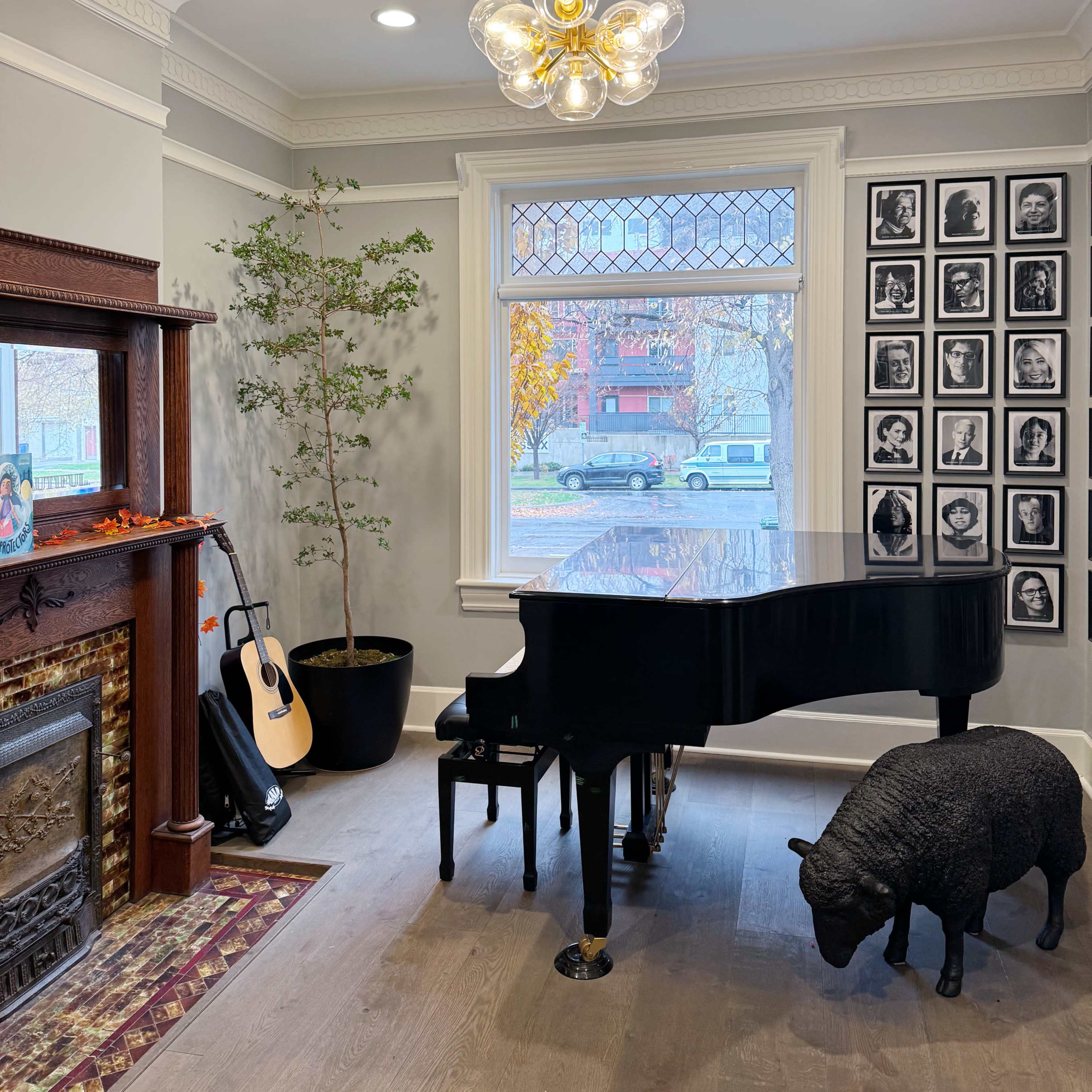 A black piano sits beside a decorative bear statue and an acoustic guitar in a cozy room featuring a chandelier and a wall of framed photographs.
