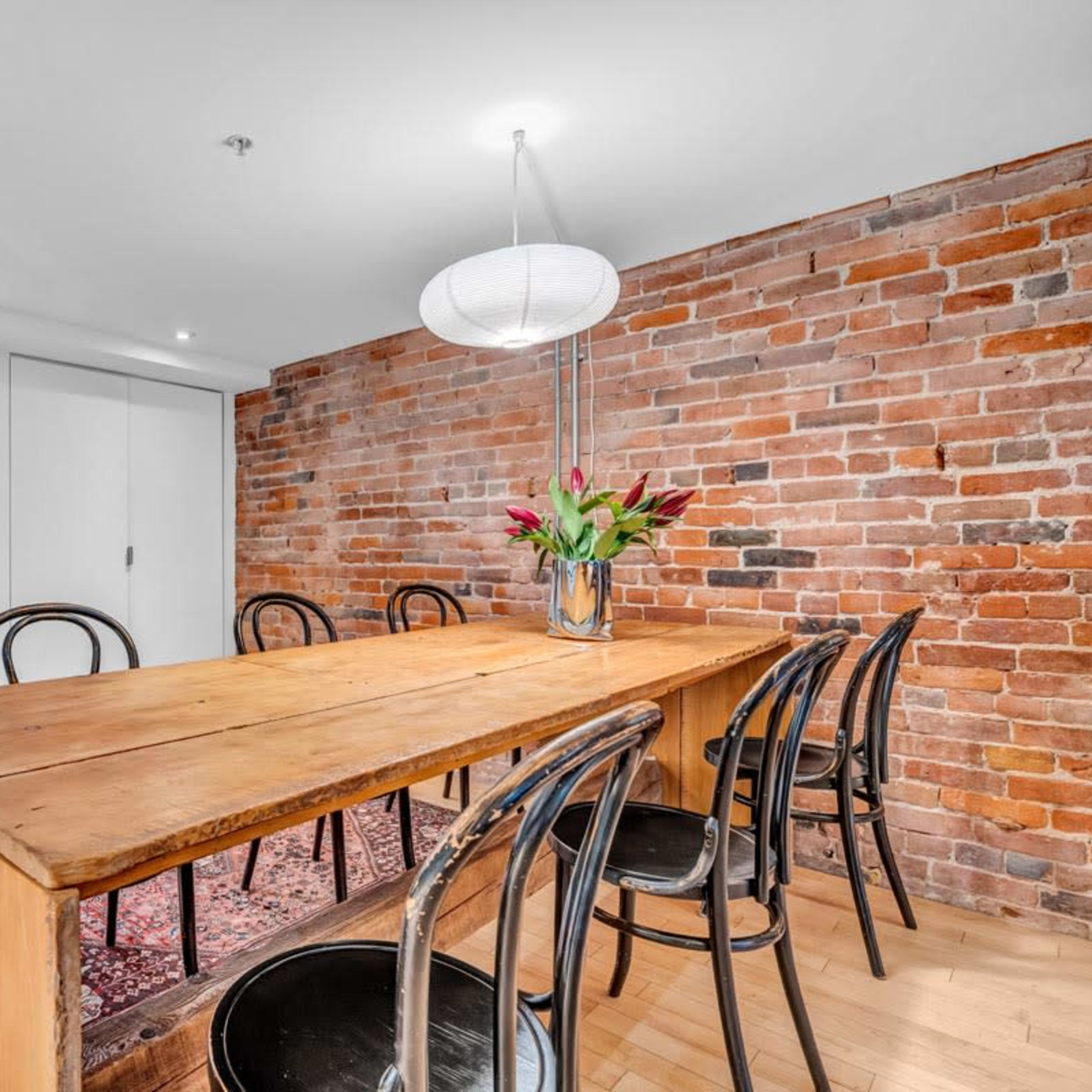 A dining area features a large wooden table with black chairs, set against a brick wall, and is illuminated by a pendant light.