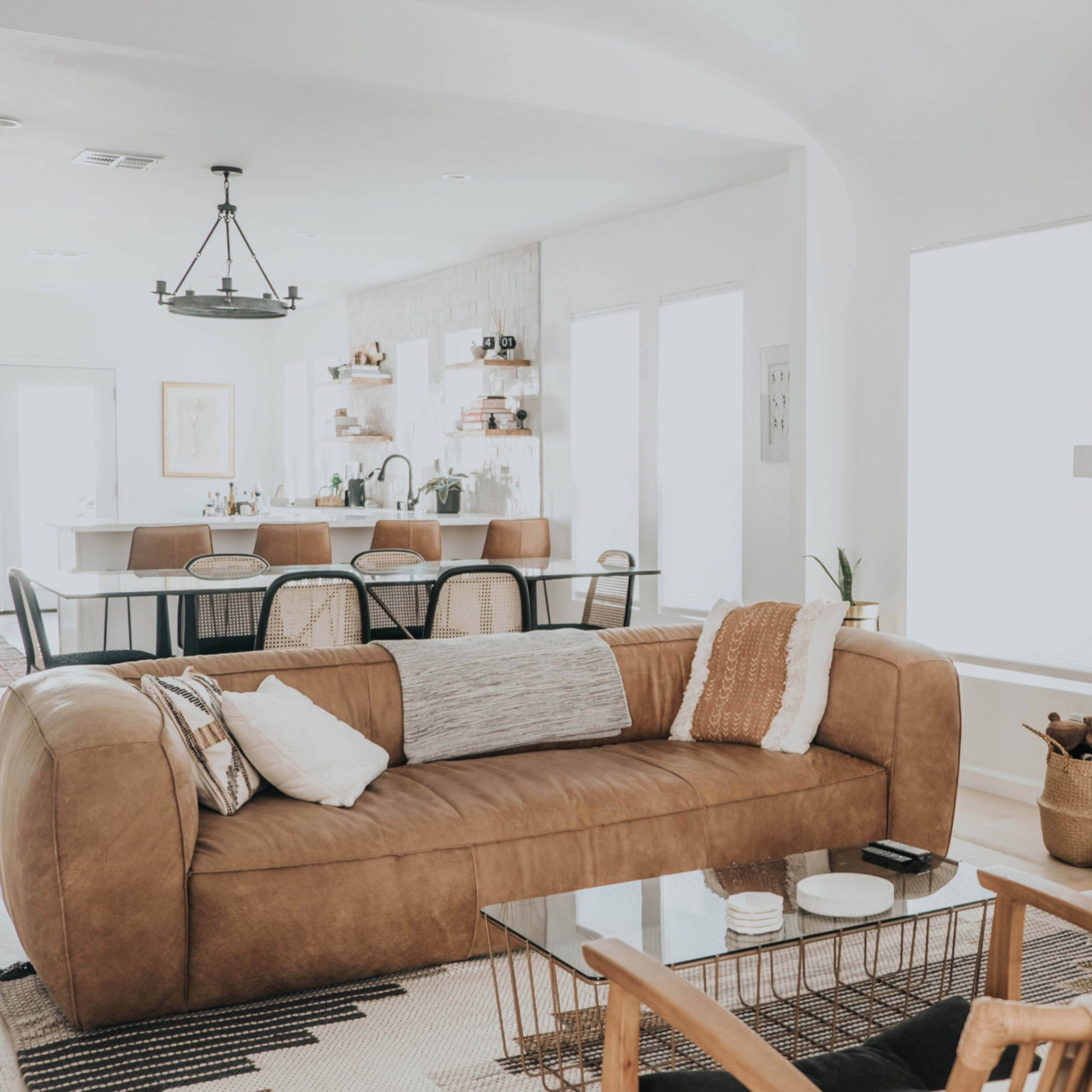 A modern living room featuring a brown leather sofa, a glass coffee table, and a fireplace with decorative elements, leading to a dining area in the background.