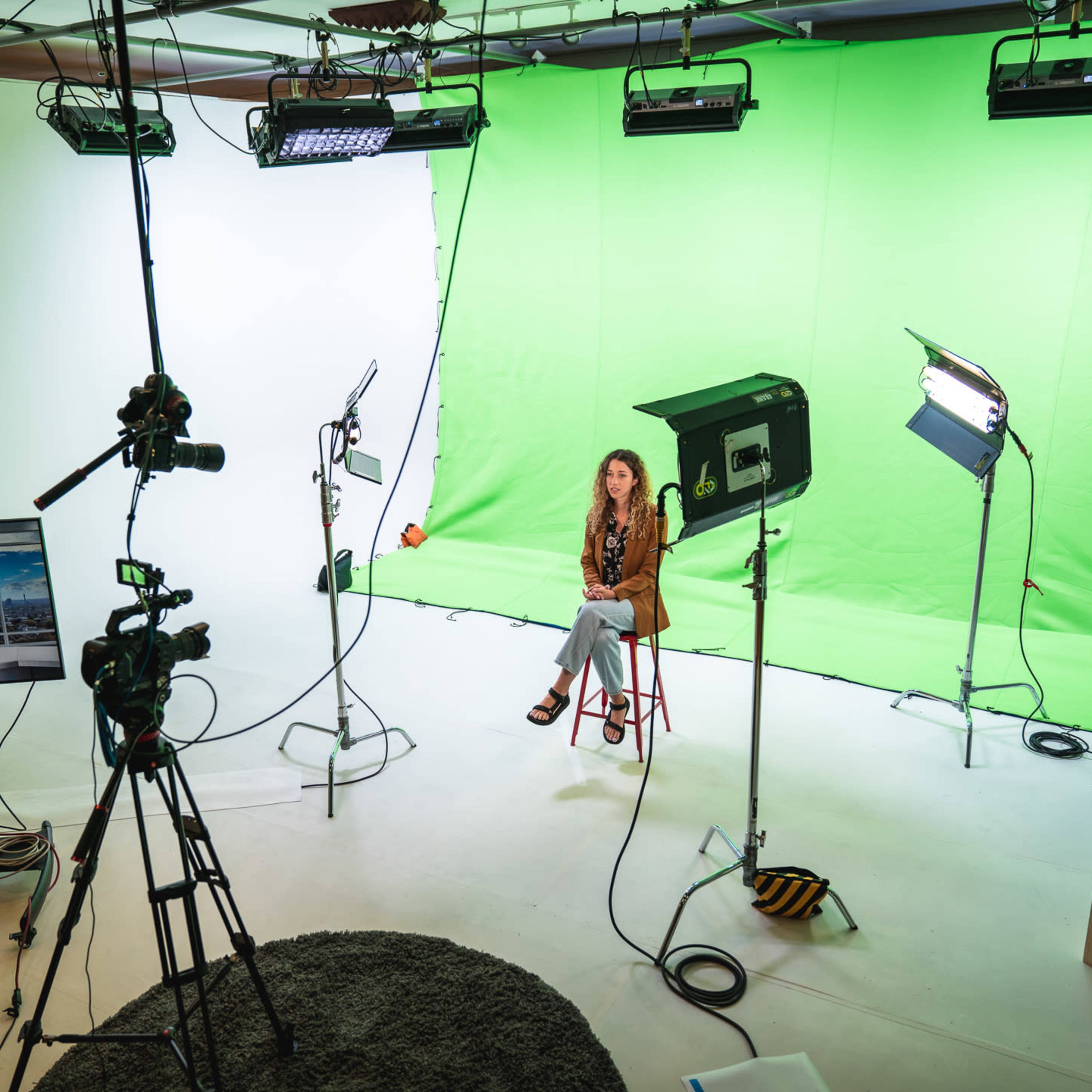 A woman is seated on a stool in a well-equipped video production studio with green and white backdrops, multiple cameras, and lighting setups.