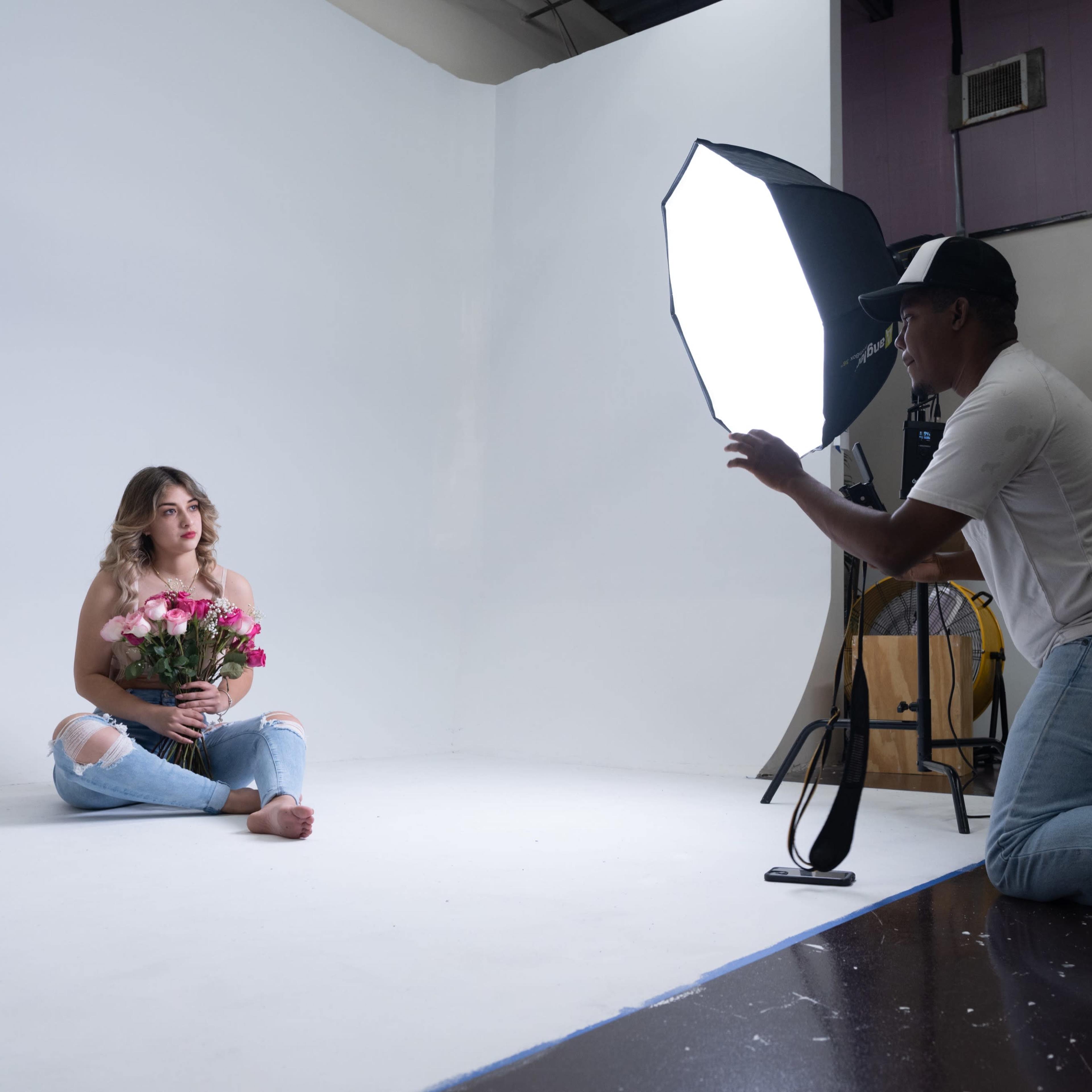 A woman sits on a studio floor holding a bouquet of flowers while a photographer kneels nearby, adjusting a light softbox.