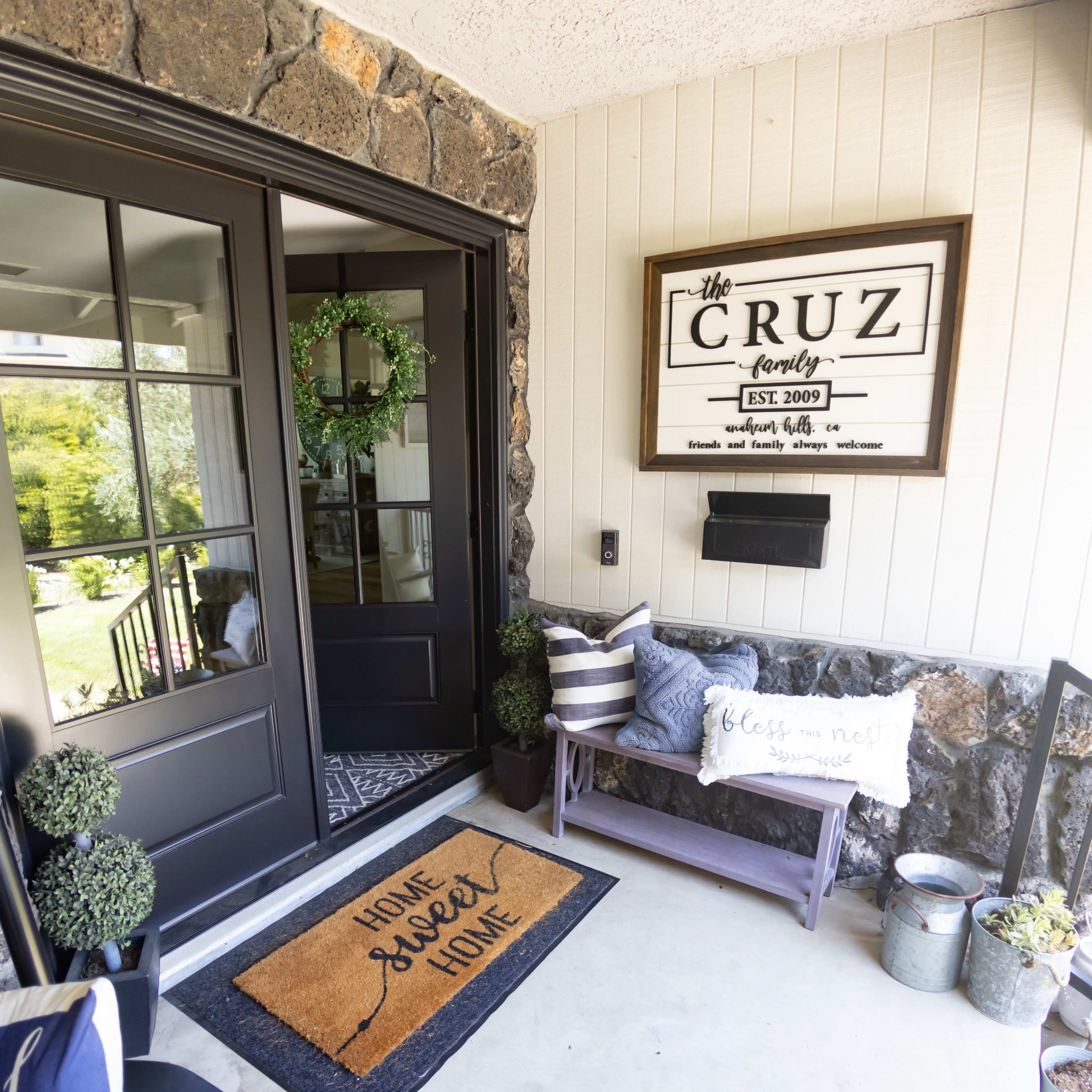 A front porch with stone walls, a dark double door, a decorative welcome mat, and various potted plants.