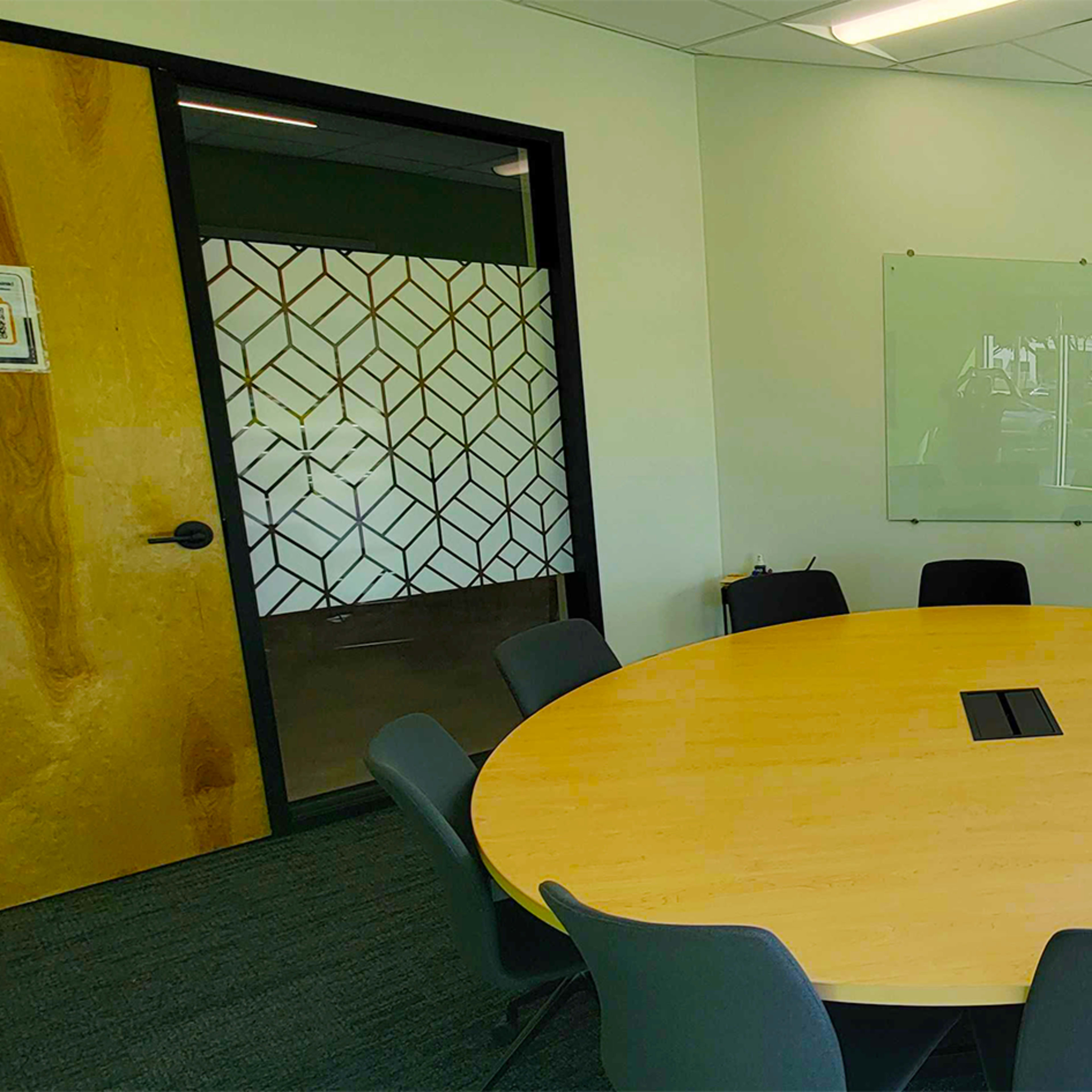 A modern conference room featuring a large circular wooden table surrounded by gray chairs, with a whiteboard on the wall and natural light coming through large windows.