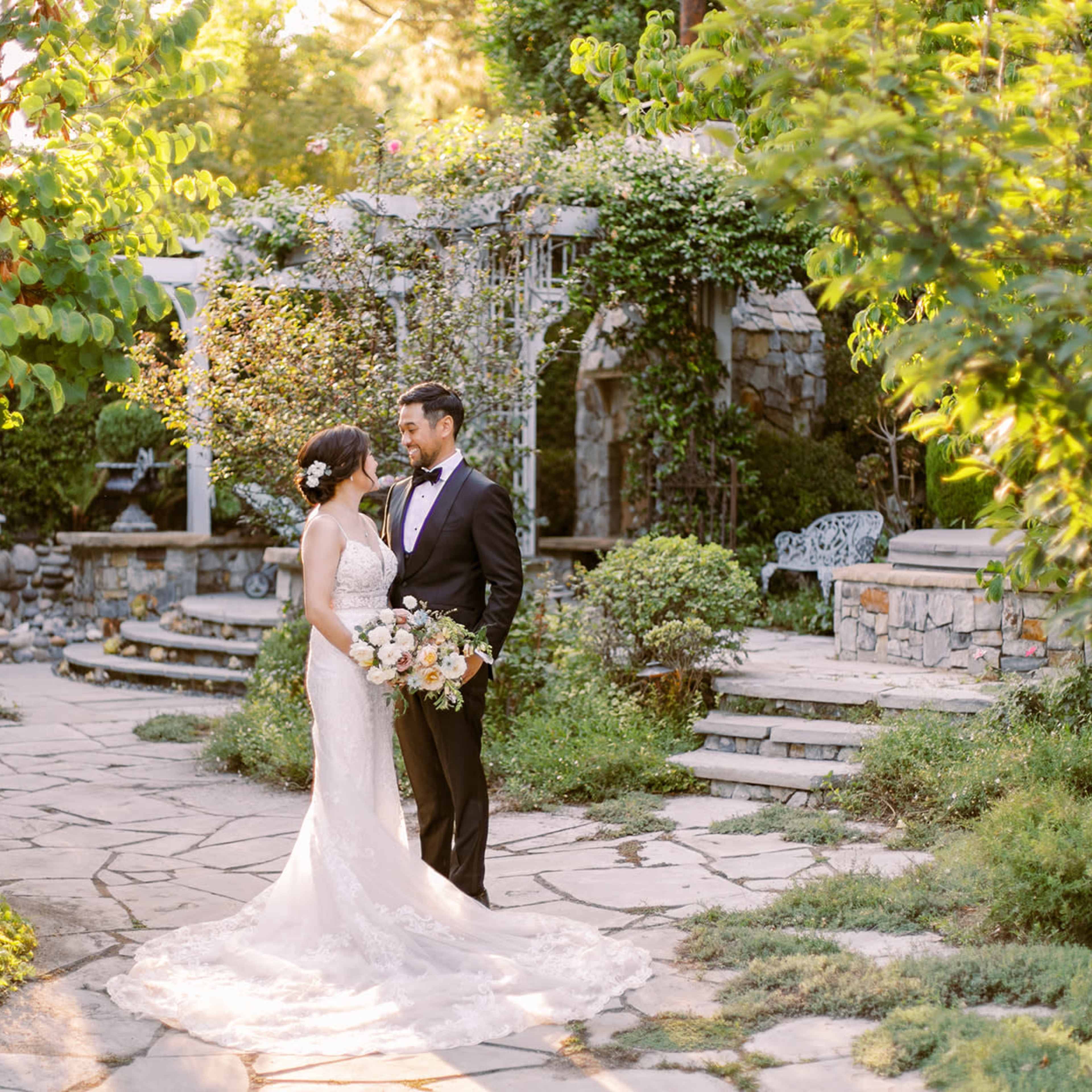 A bride and groom stand together in a stone-walled garden surrounded by greenery and floral arrangements.