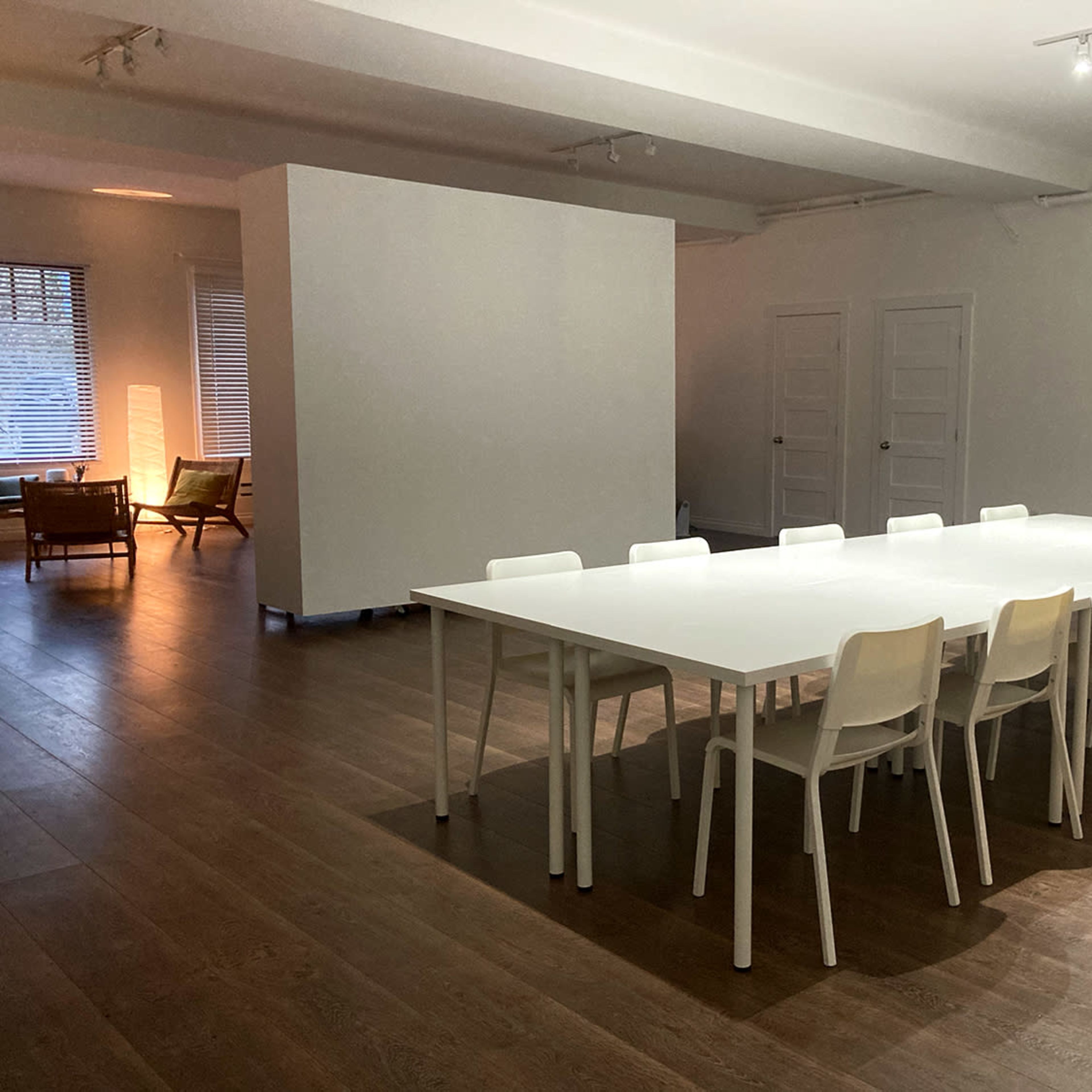 A large, empty meeting room features a long white table surrounded by ten white chairs, with wooden flooring and a partition in the background.