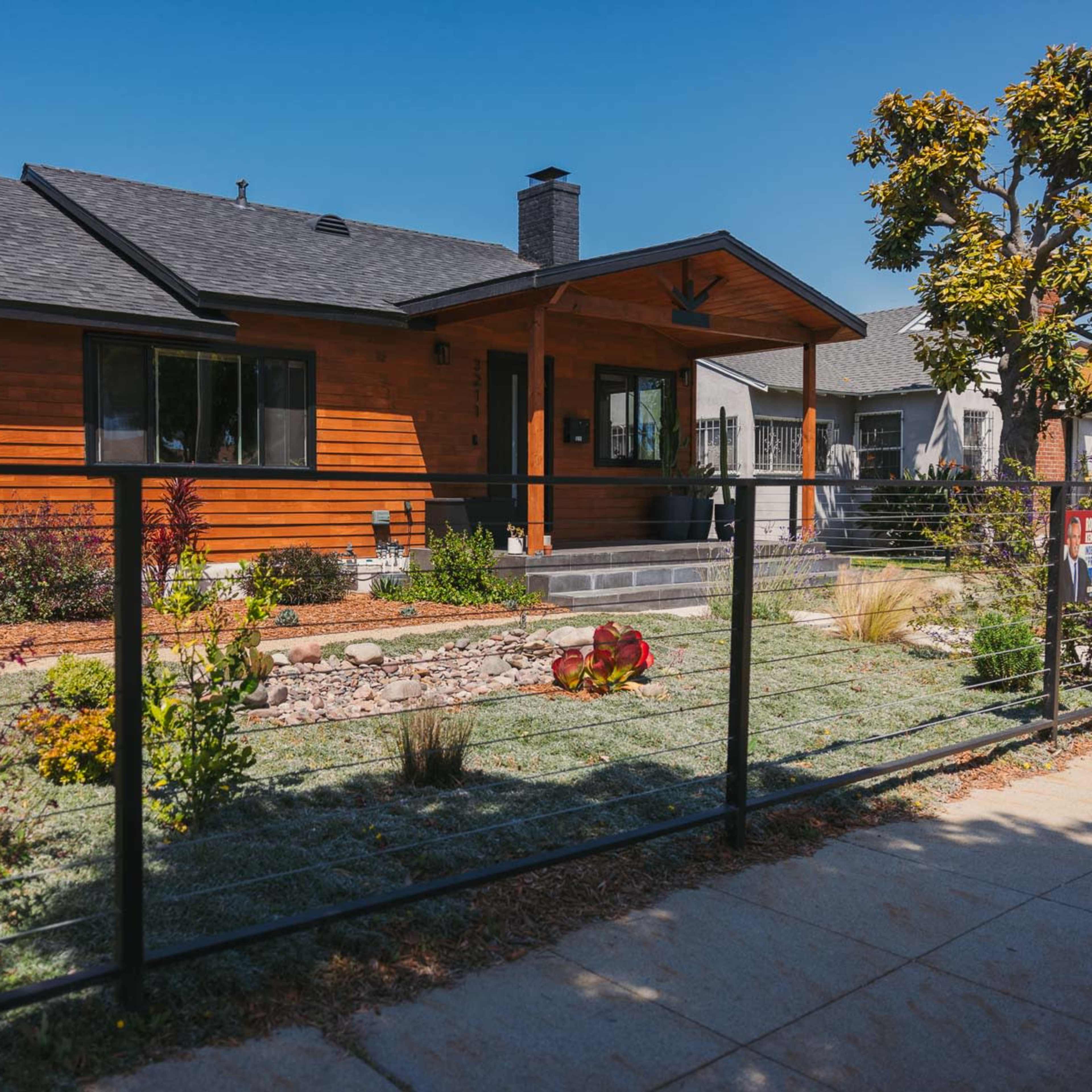 A modern single-story house with a wooden facade and a landscaped front yard is separated from the sidewalk by a black metal fence.