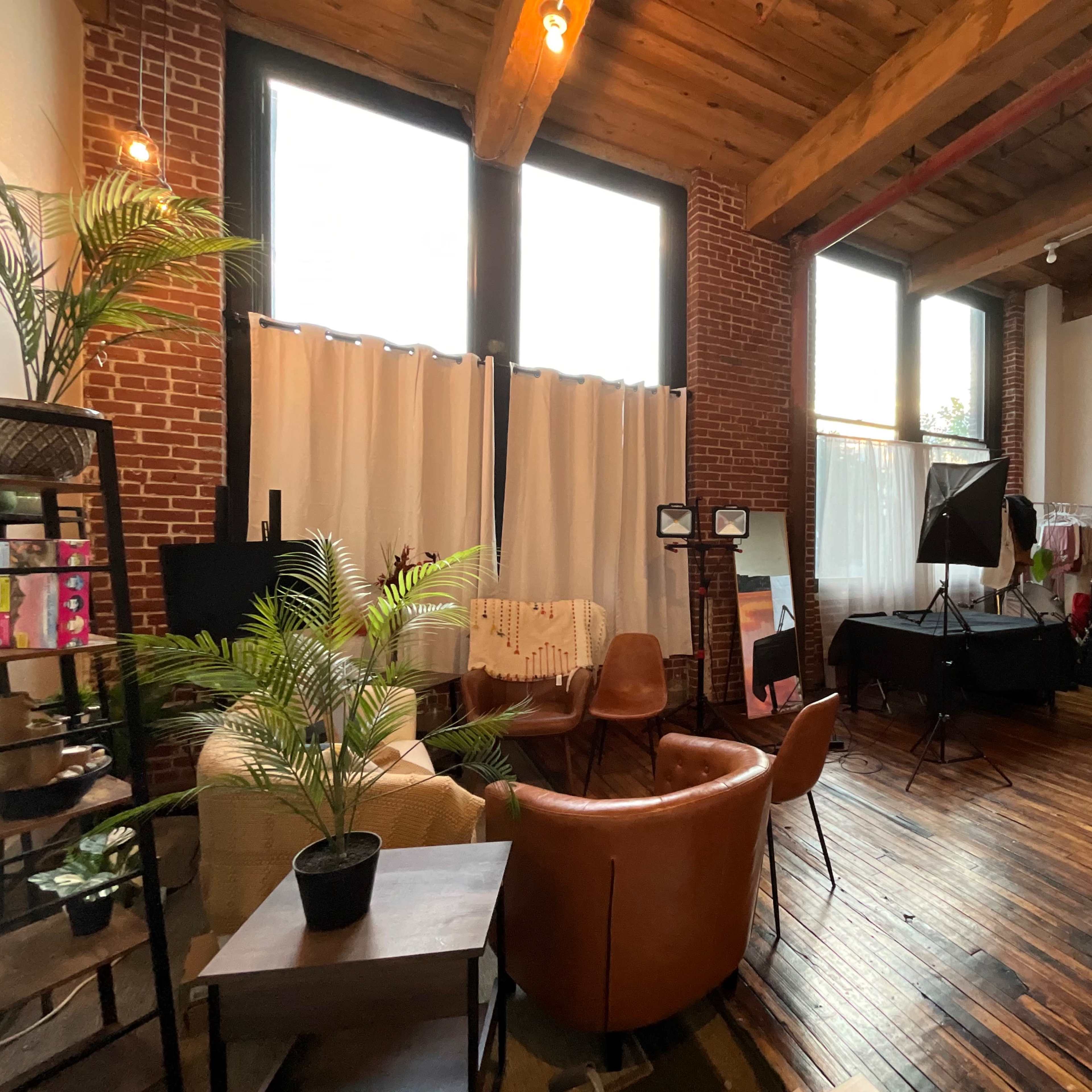 A spacious studio with brick walls, large windows, and various seating arrangements, surrounded by photography equipment and plants.