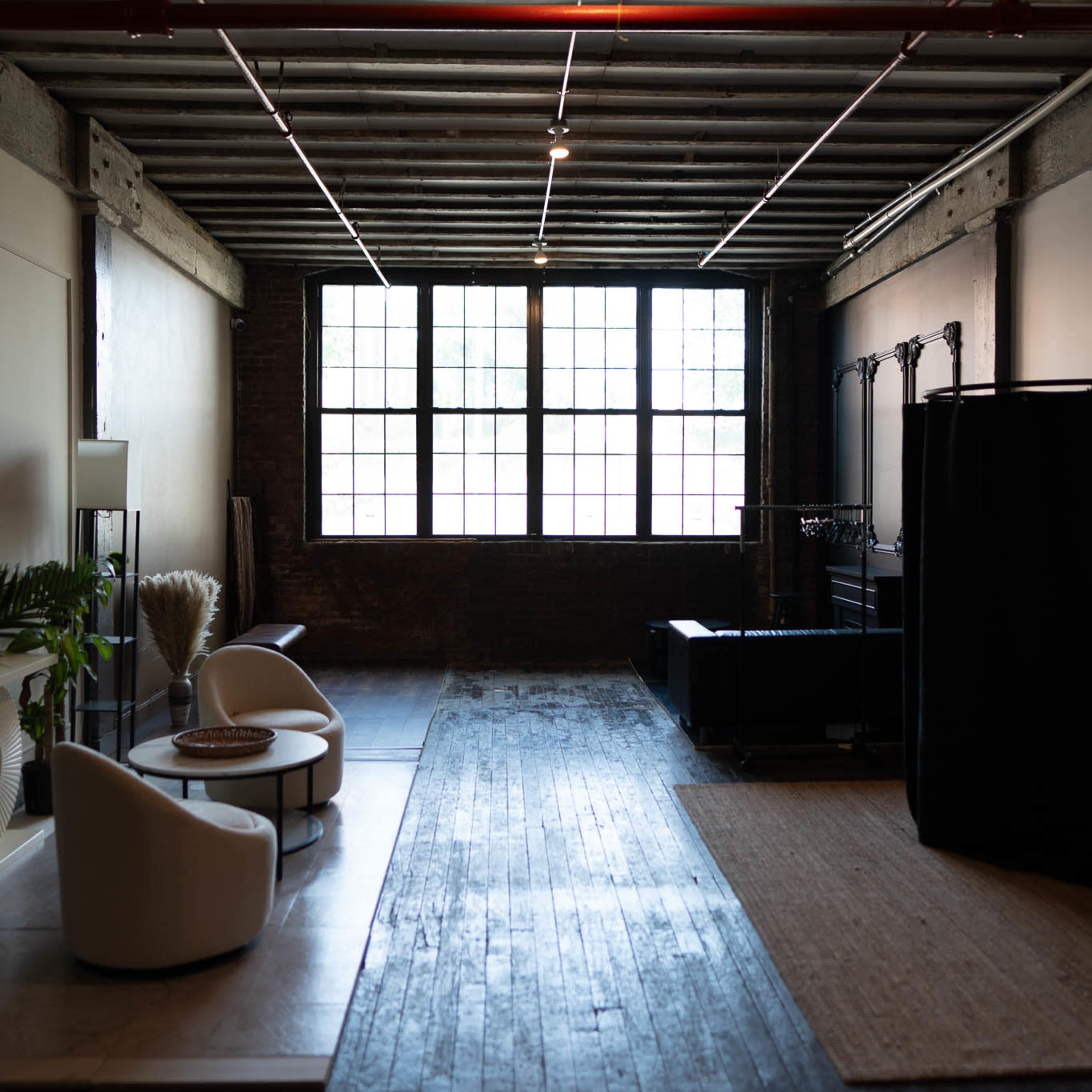 A spacious industrial-style interior with wooden floors, large windows, and minimal furnishings including two white chairs and a small round table.