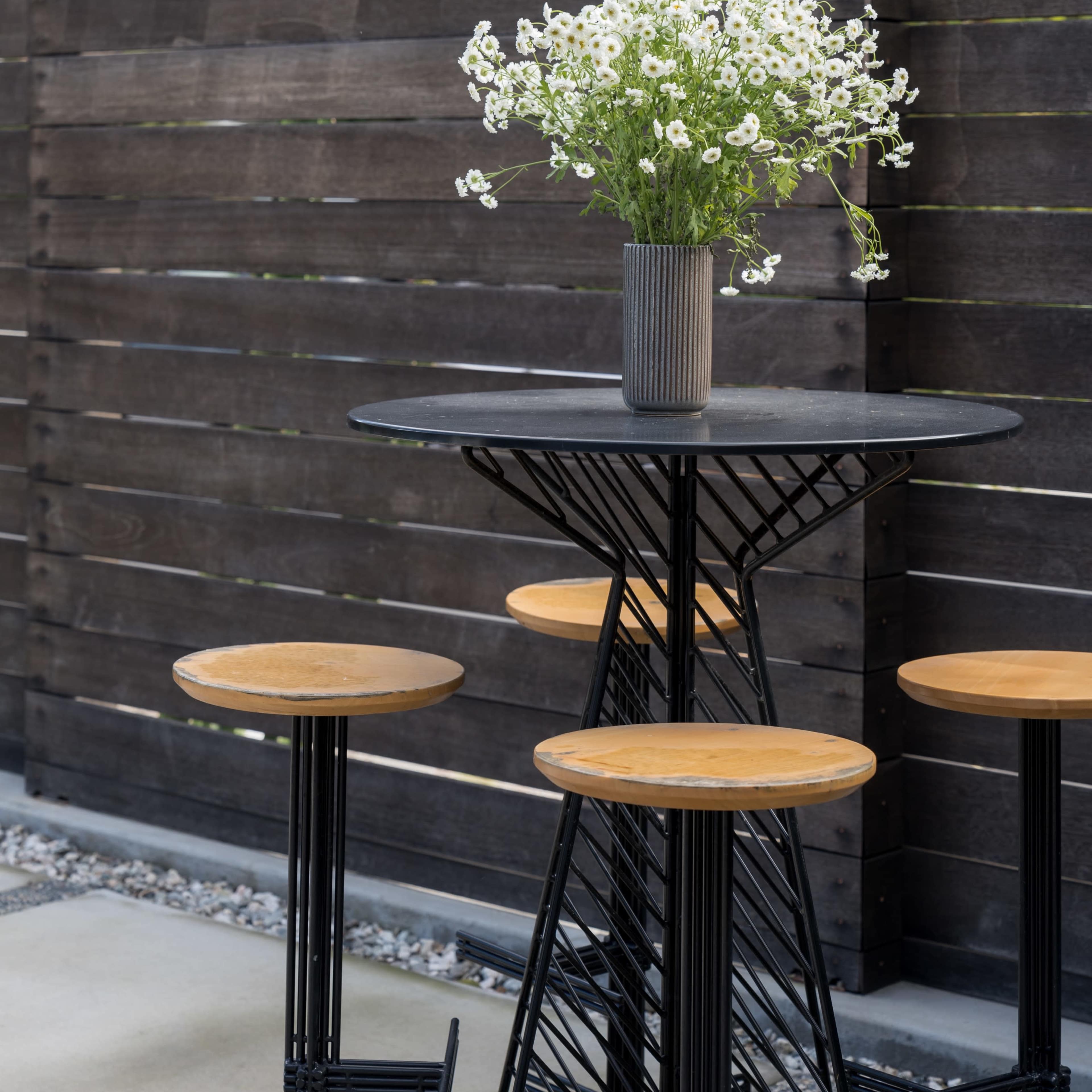 A black metal table with wooden stools is set against a backdrop of a wooden fence and a tree, featuring a vase of white flowers on the table.