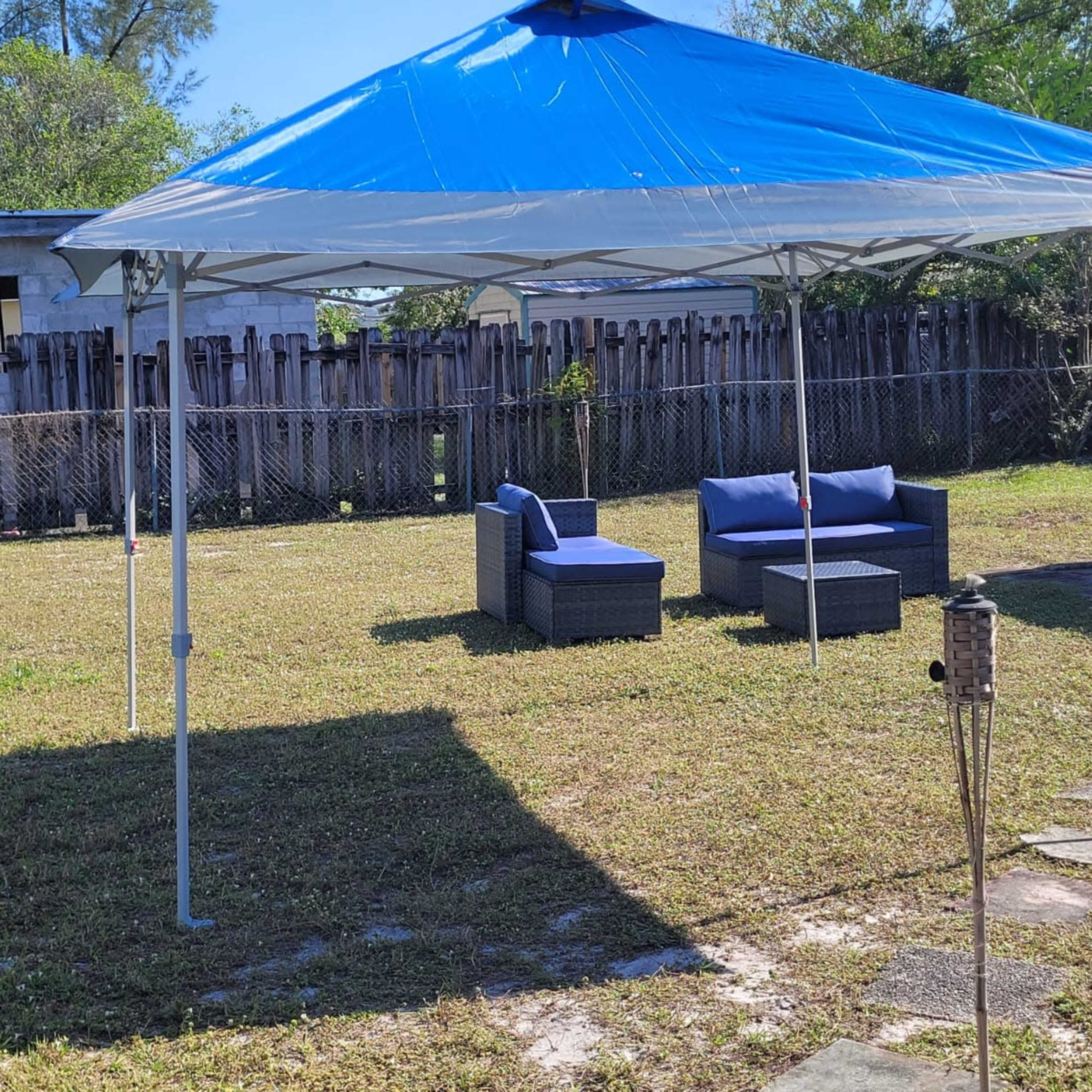 A blue and white canopy tent is set up in a grassy area with two cushioned chairs and a table beneath it.