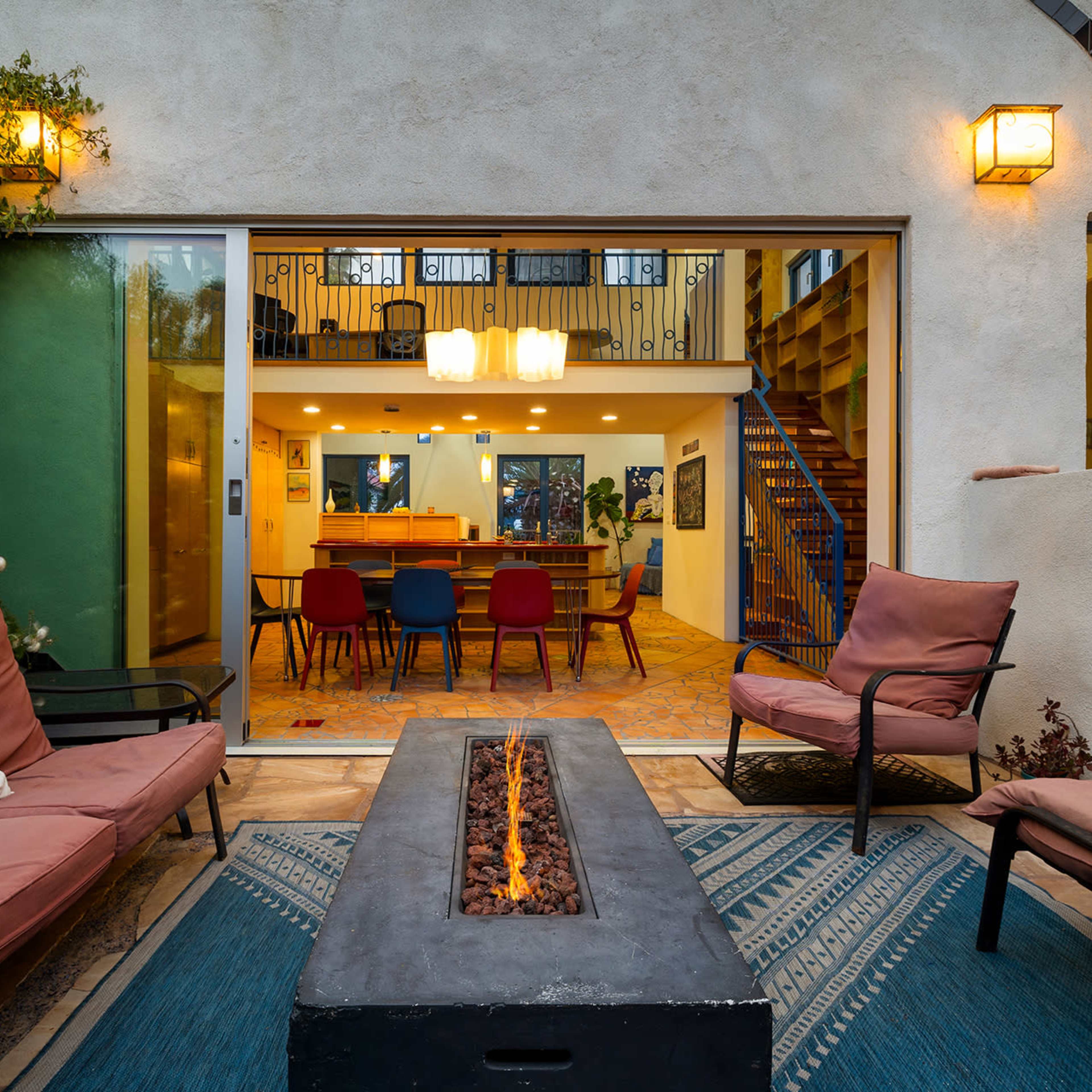 A cozy outdoor seating area features two chairs and a fire pit, with large glass doors opening to a brightly lit interior dining space.