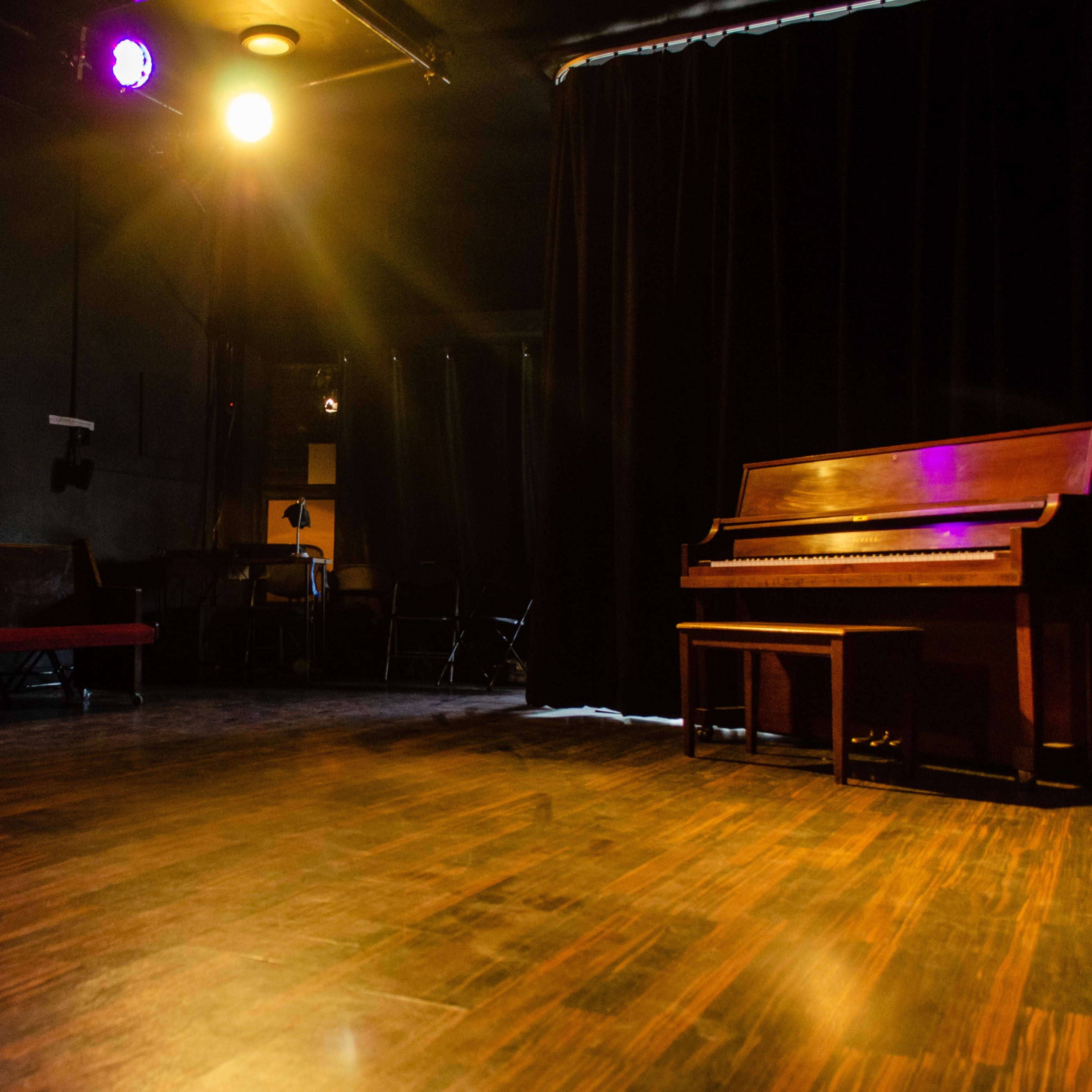 A dimly lit stage features a wooden piano and a few chairs in a small theater setting.