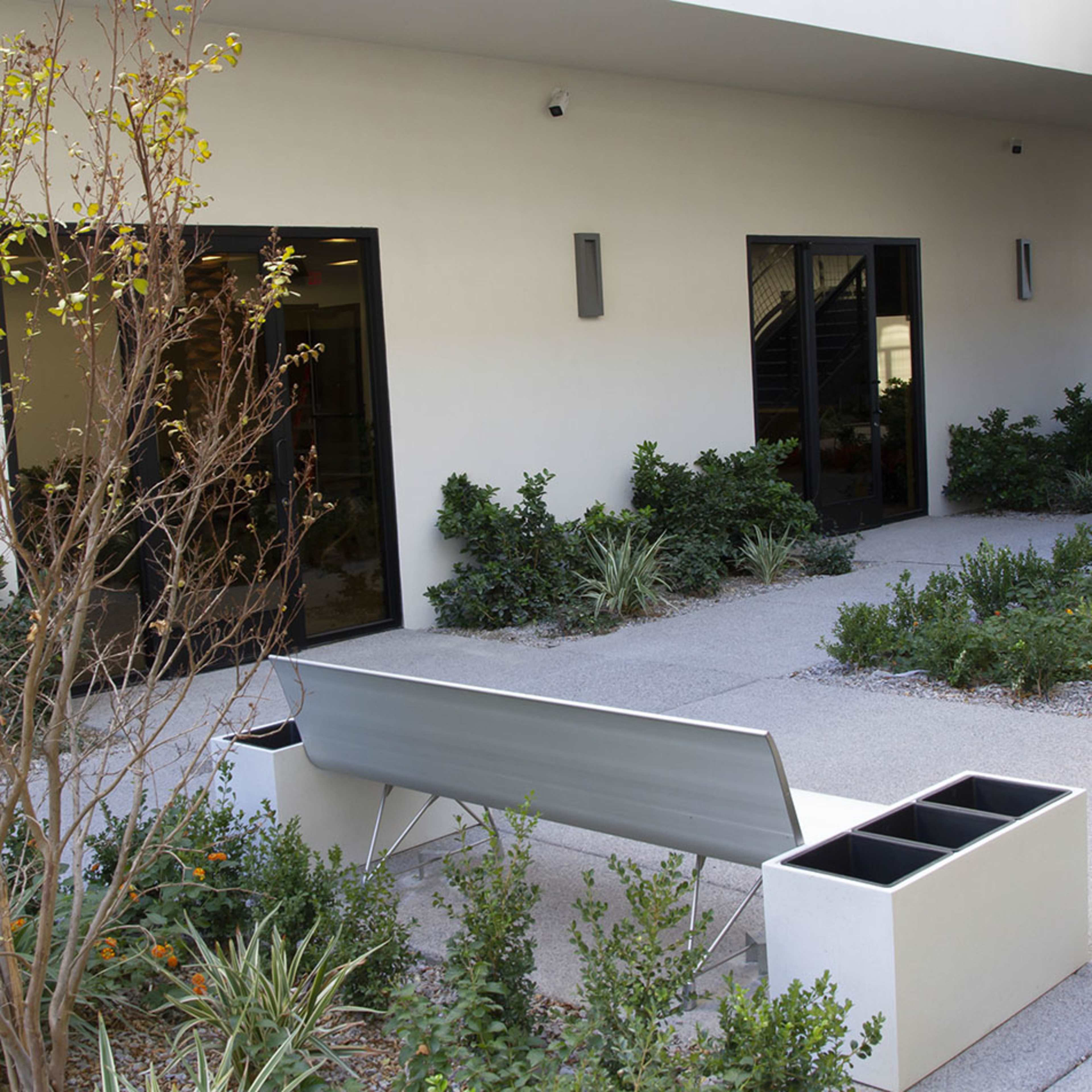 A modern bench is positioned near two planter boxes on a landscaped pathway adjacent to a building.