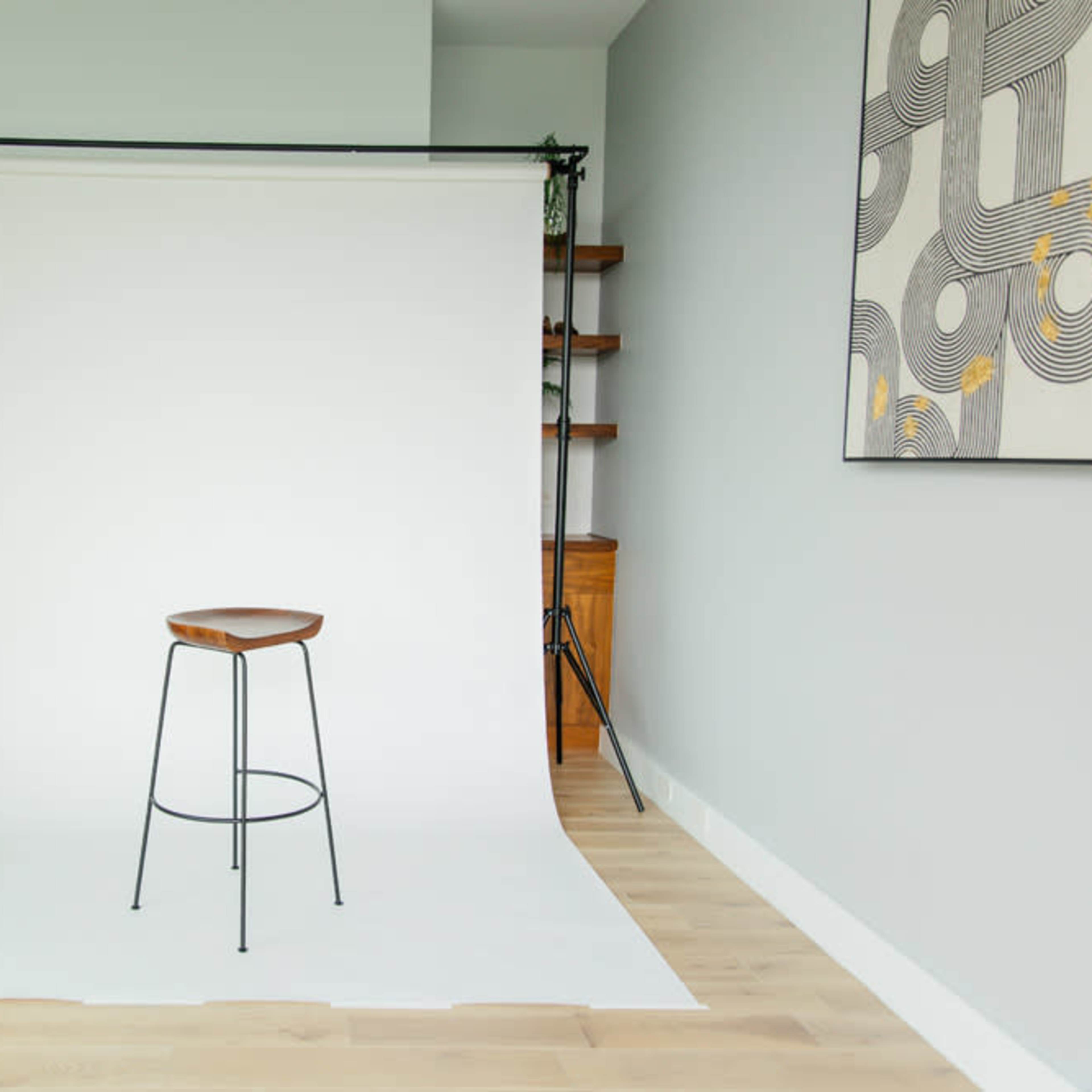 A minimalistic studio setup with a single wooden stool in front of a white backdrop and light-colored wooden flooring.