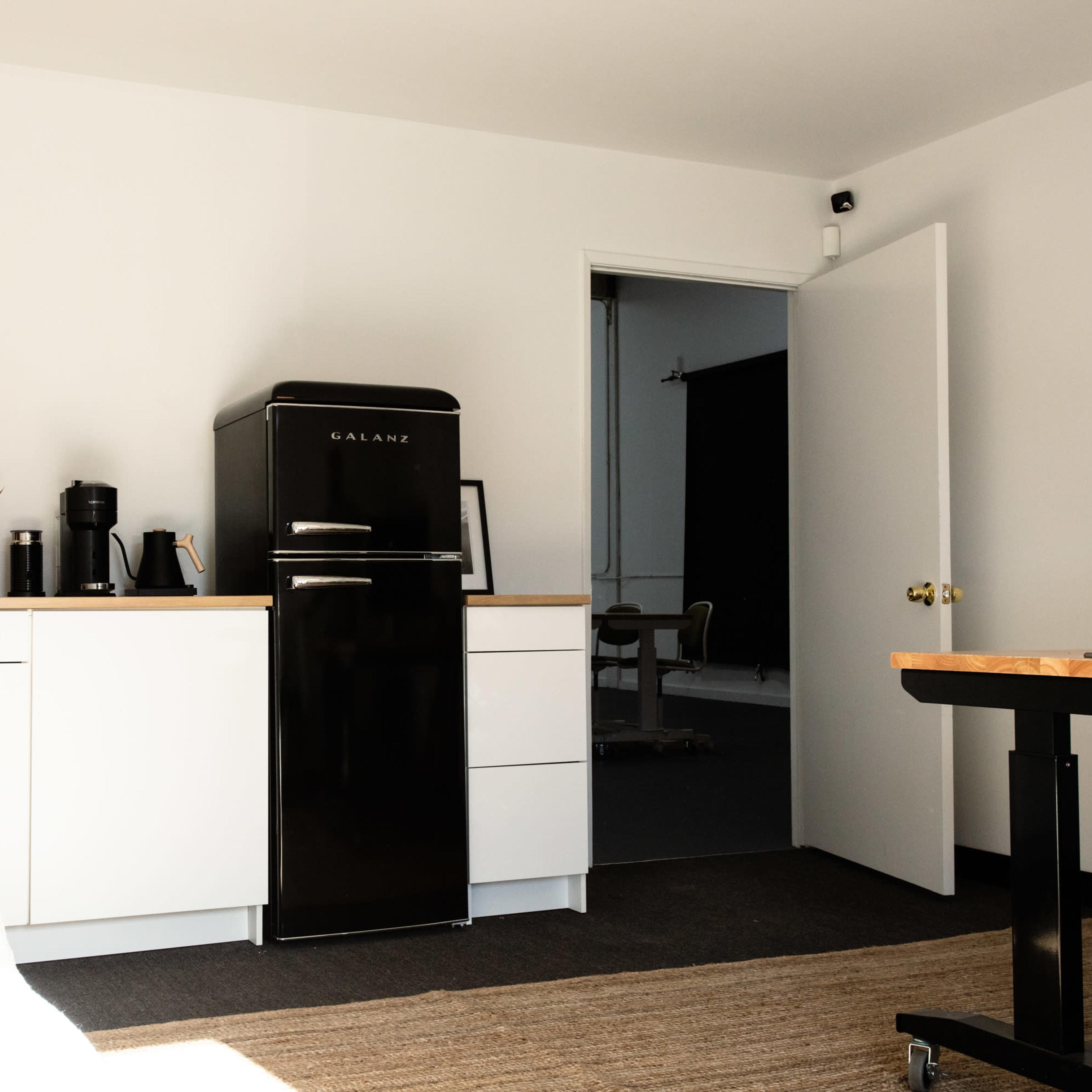 A modern office space featuring a black refrigerator, white cabinetry, a coffee maker, and a doorway leading to an adjacent room.