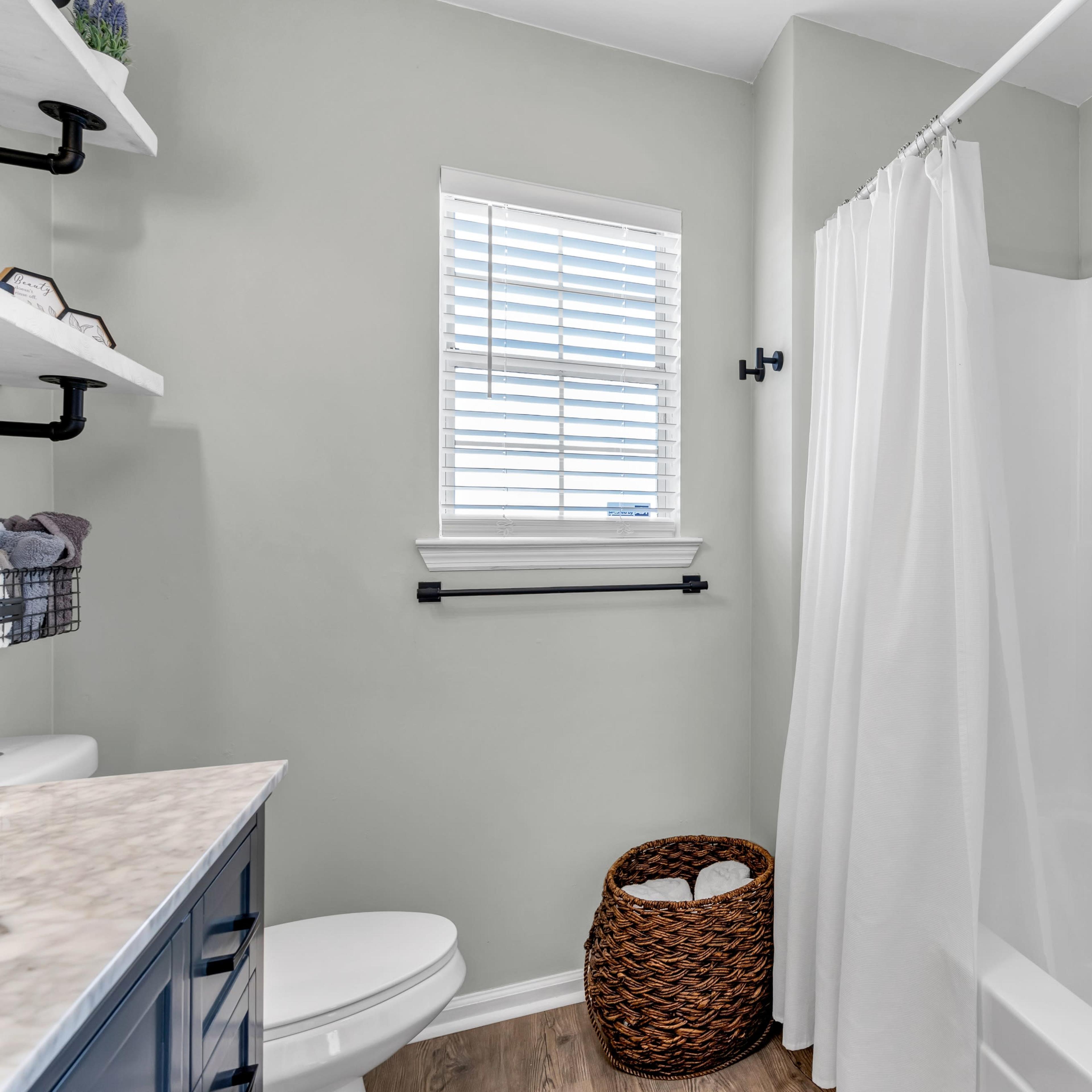 A bathroom featuring a white bathtub with a shower curtain, a toilet, and a woven basket for storage, all set against green walls and a window with blinds.