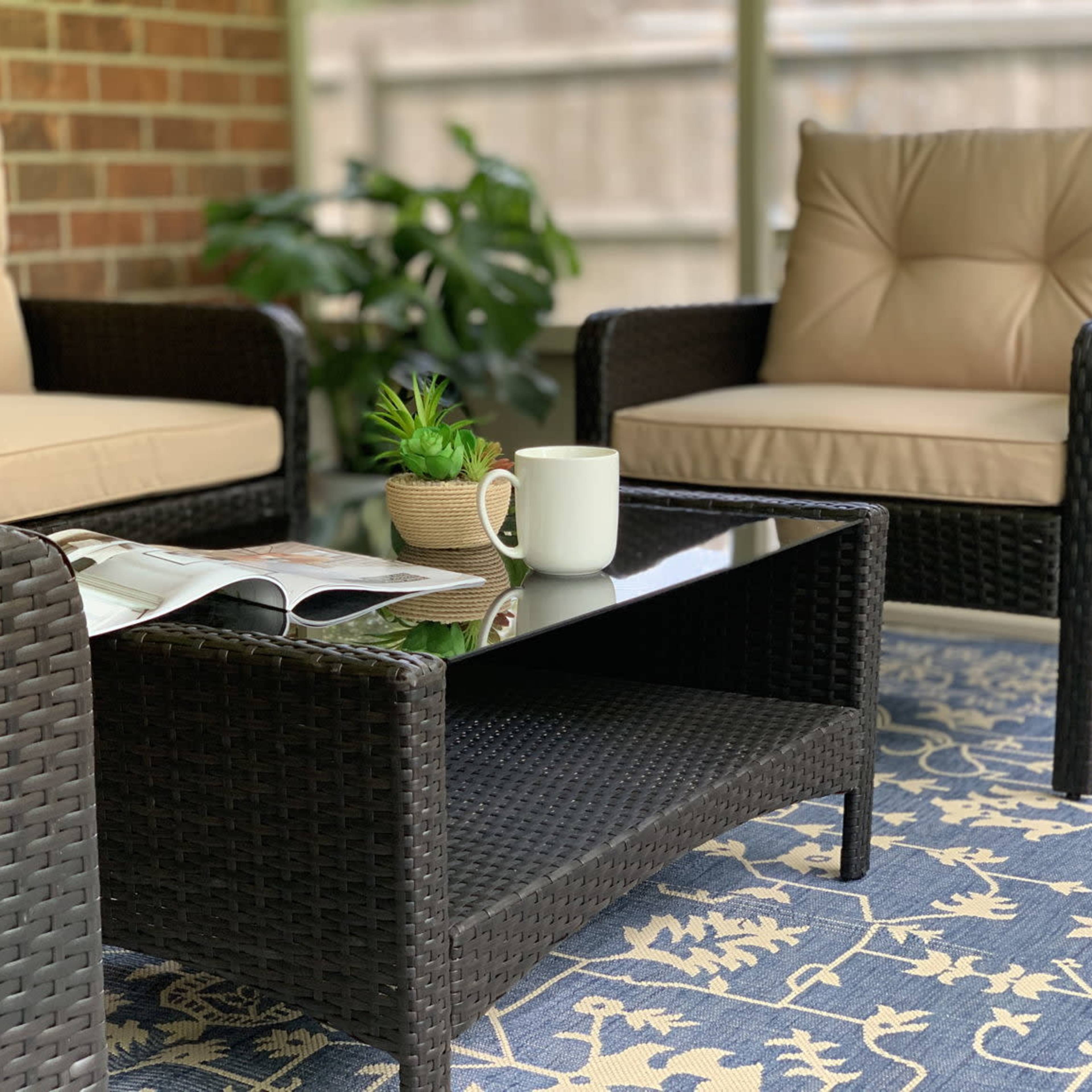 A cozy seating area with two wicker chairs and a coffee table, set on a patterned rug, with a plant and a magazine on the table.