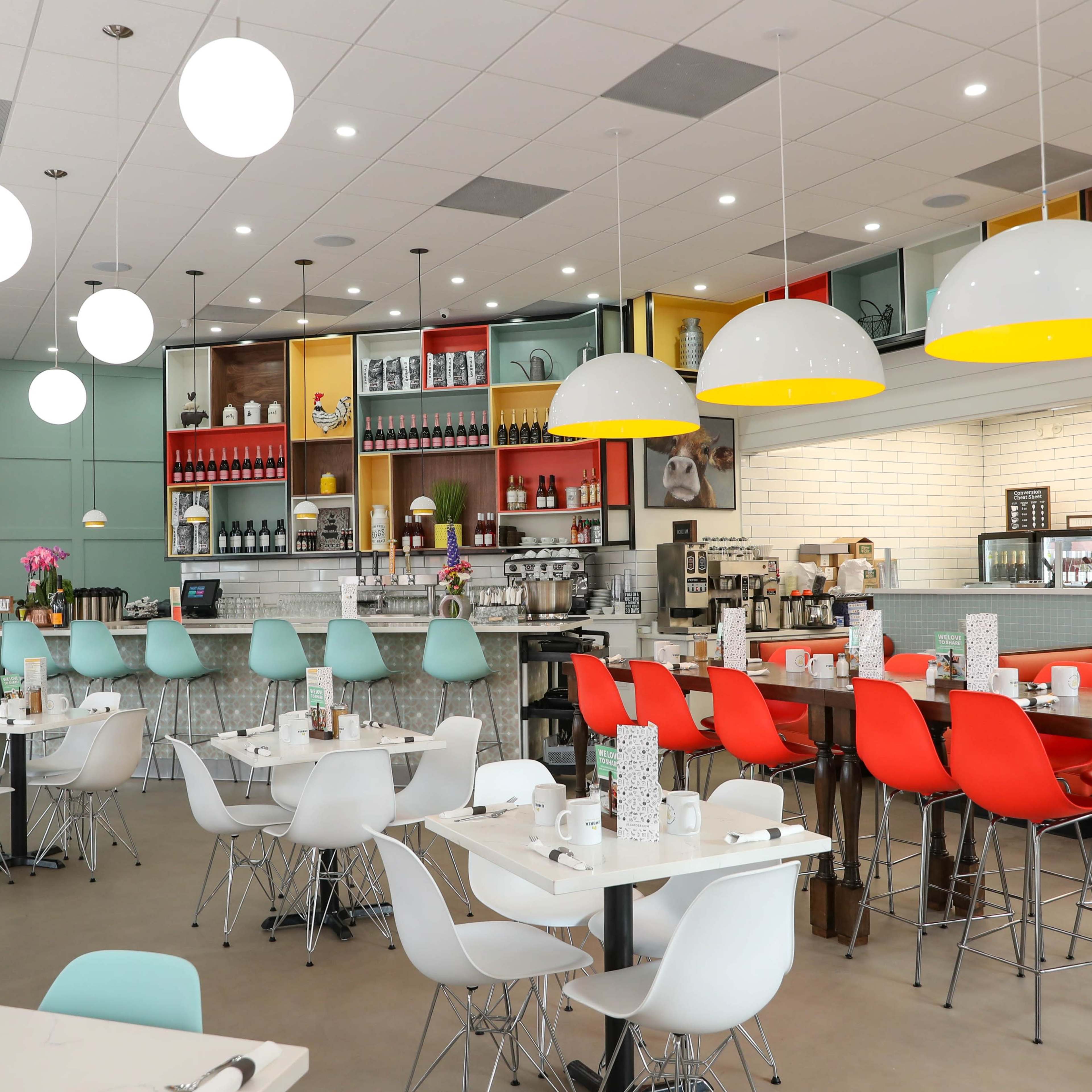 A modern restaurant features colorful chairs and pendant lighting, with tables set for dining and a well-stocked bar area in the background.
