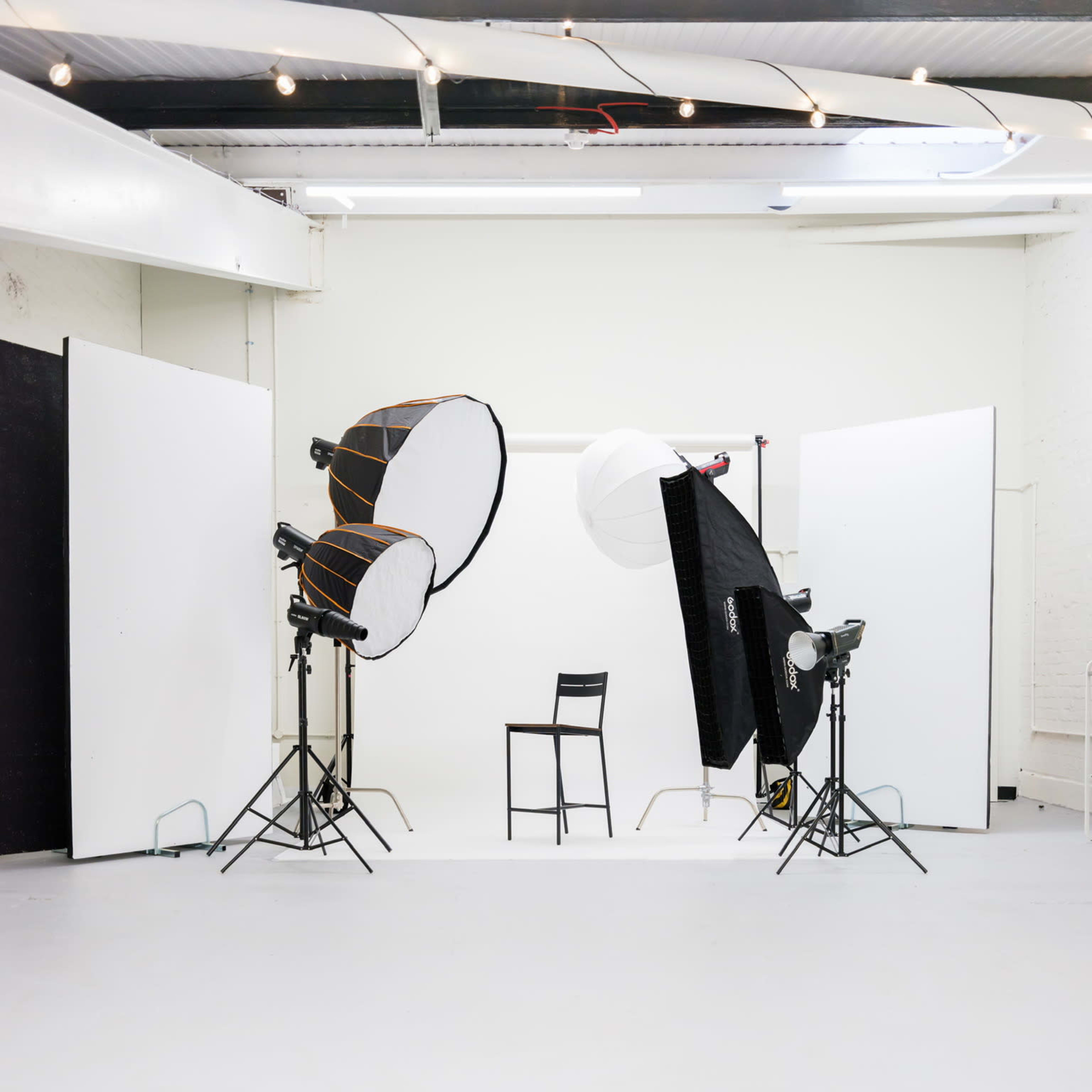 A photography studio is set up with two lighting setups and a single chair positioned in the center against a white backdrop.