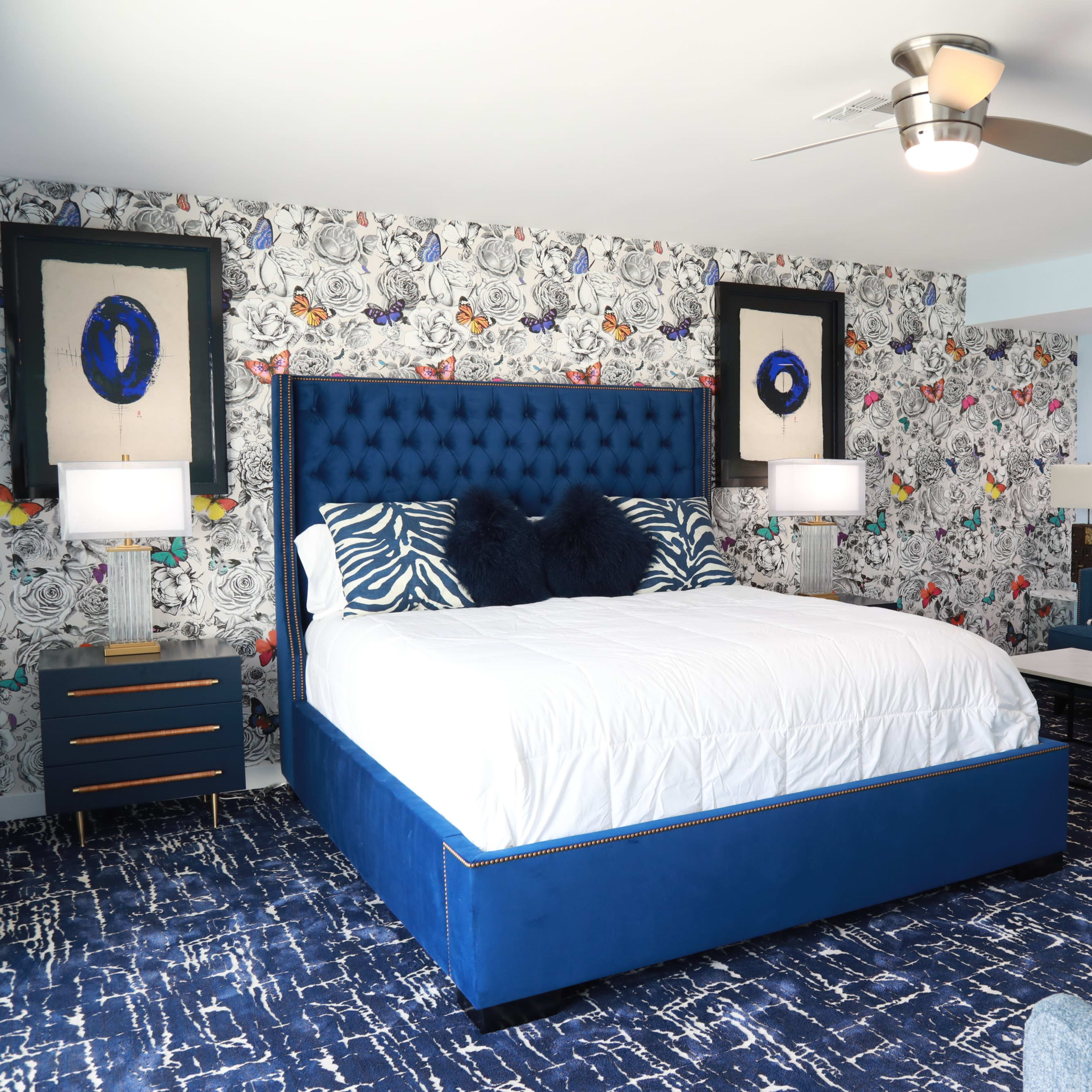 A modern bedroom featuring a blue tufted bed with white bedding, a floral wallpaper, and a seating area with a couch and coffee table.