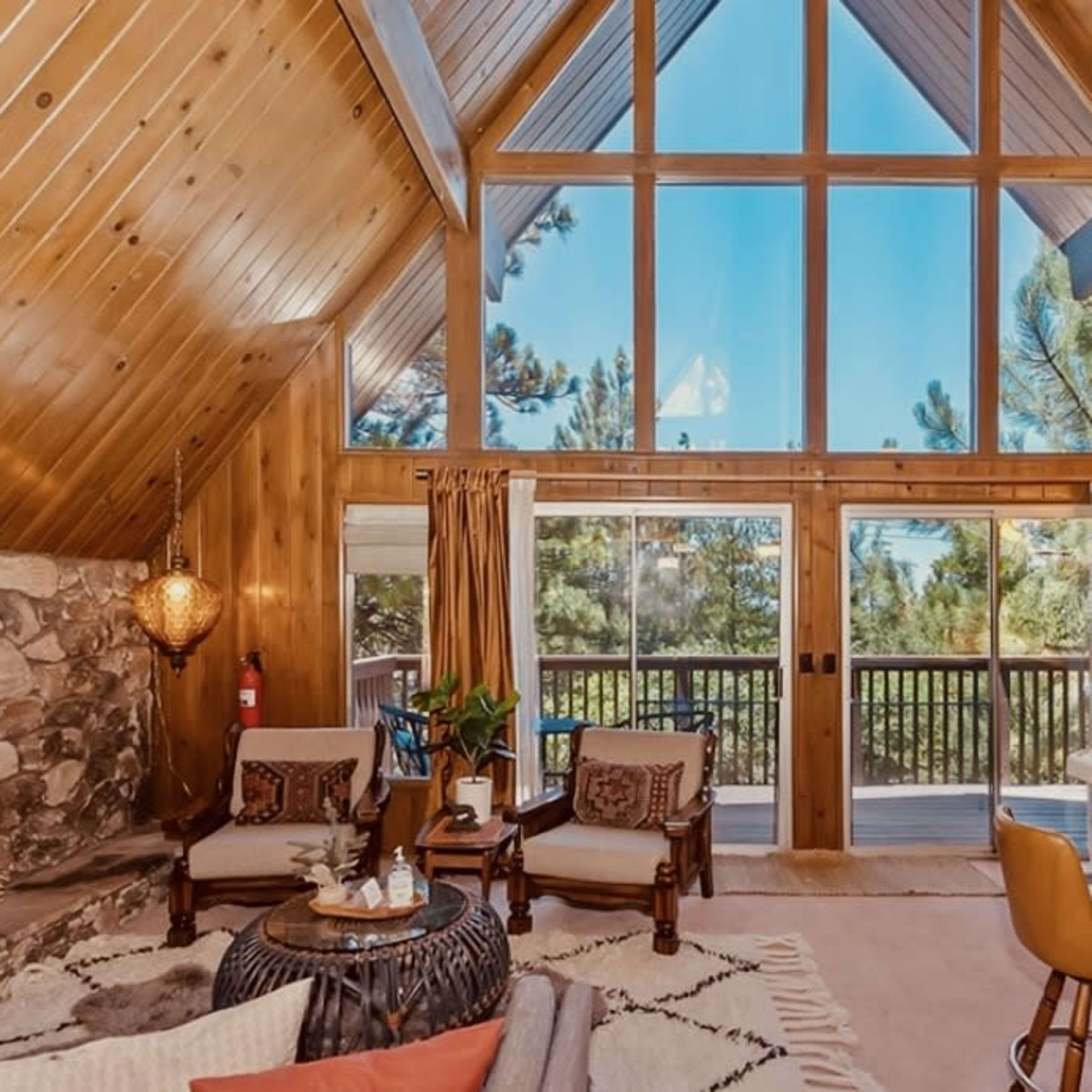 A rustic cabin interior features wooden paneling, large windows, and a stone fireplace with seating arrangements and a dining area.