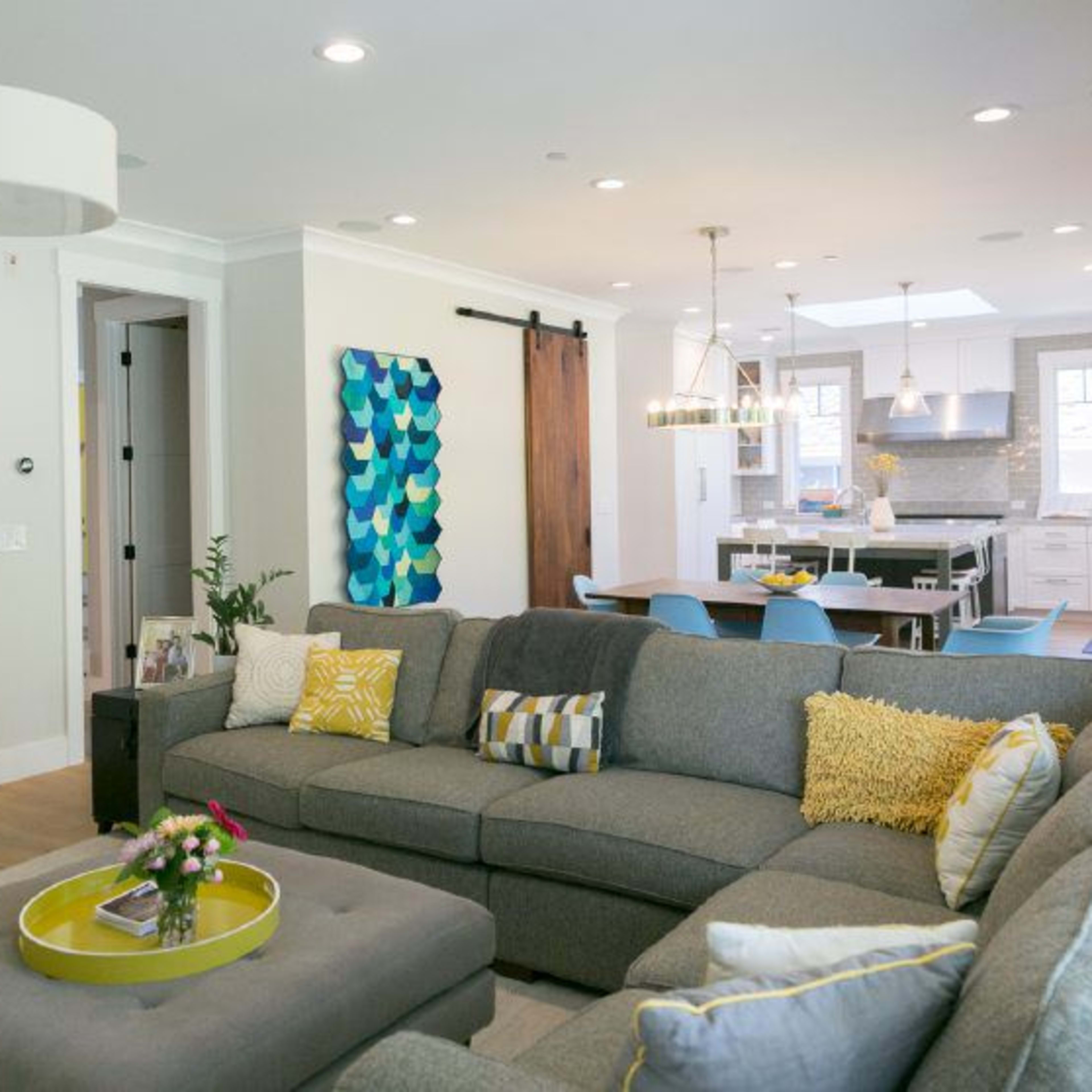 A modern living room with a large sectional sofa, a round coffee table, and artwork on the walls, leading into a brightly lit kitchen space.