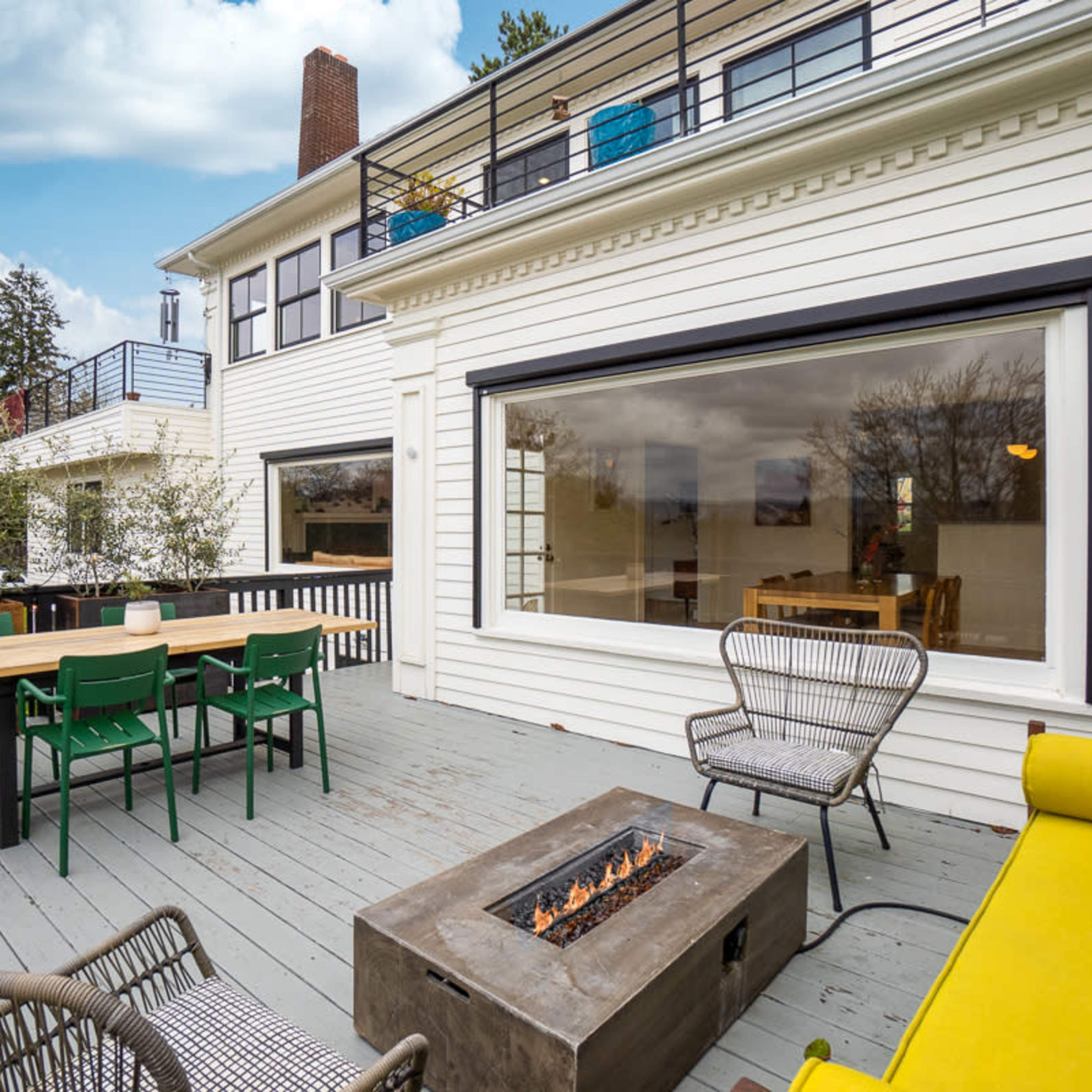 A spacious wooden deck with a fire pit, surrounded by outdoor seating and a dining table, adjacent to a modern house.