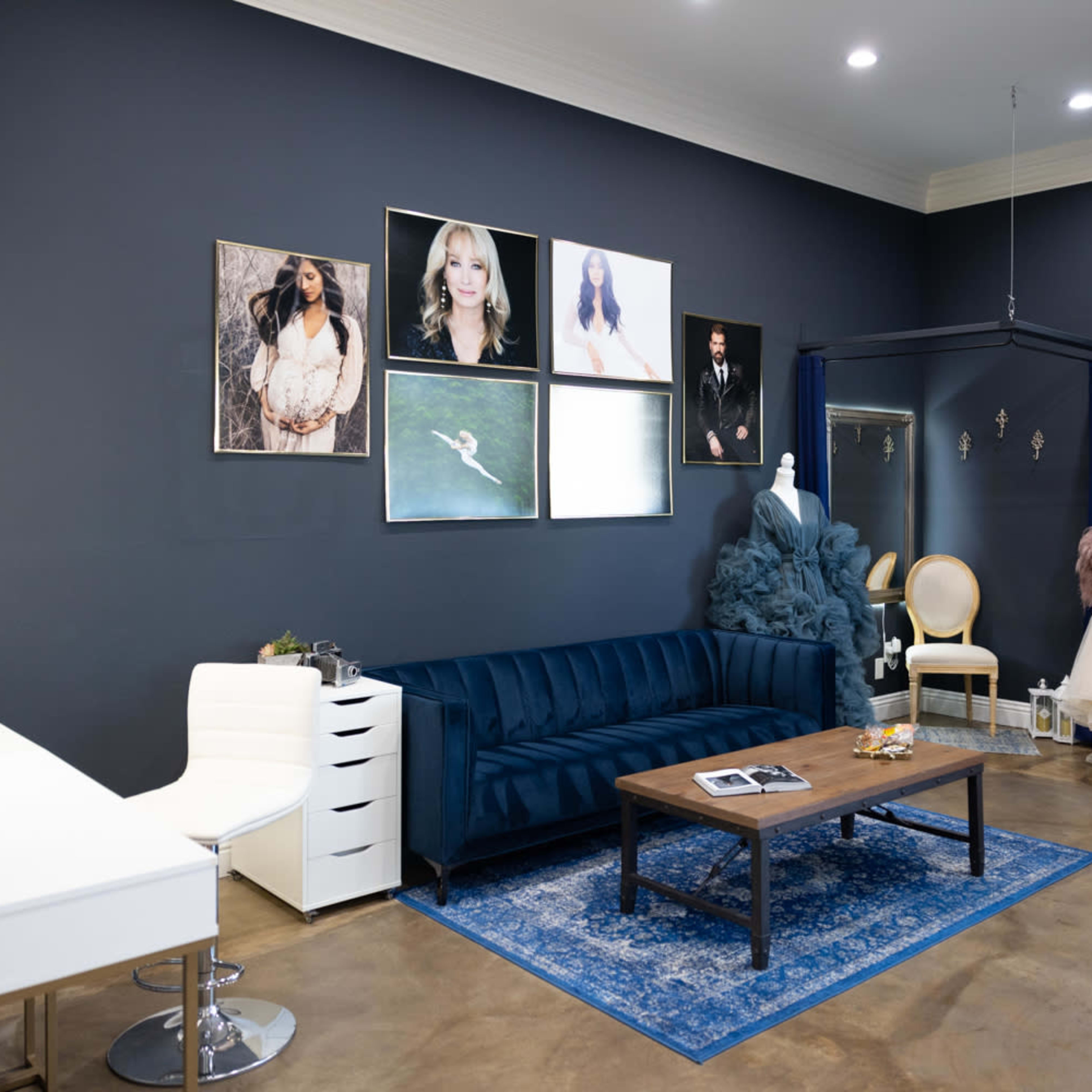A stylish studio featuring a dark blue wall, a blue velvet sofa, a wooden coffee table, and various framed photographs, along with several elegant dresses displayed on mannequins.