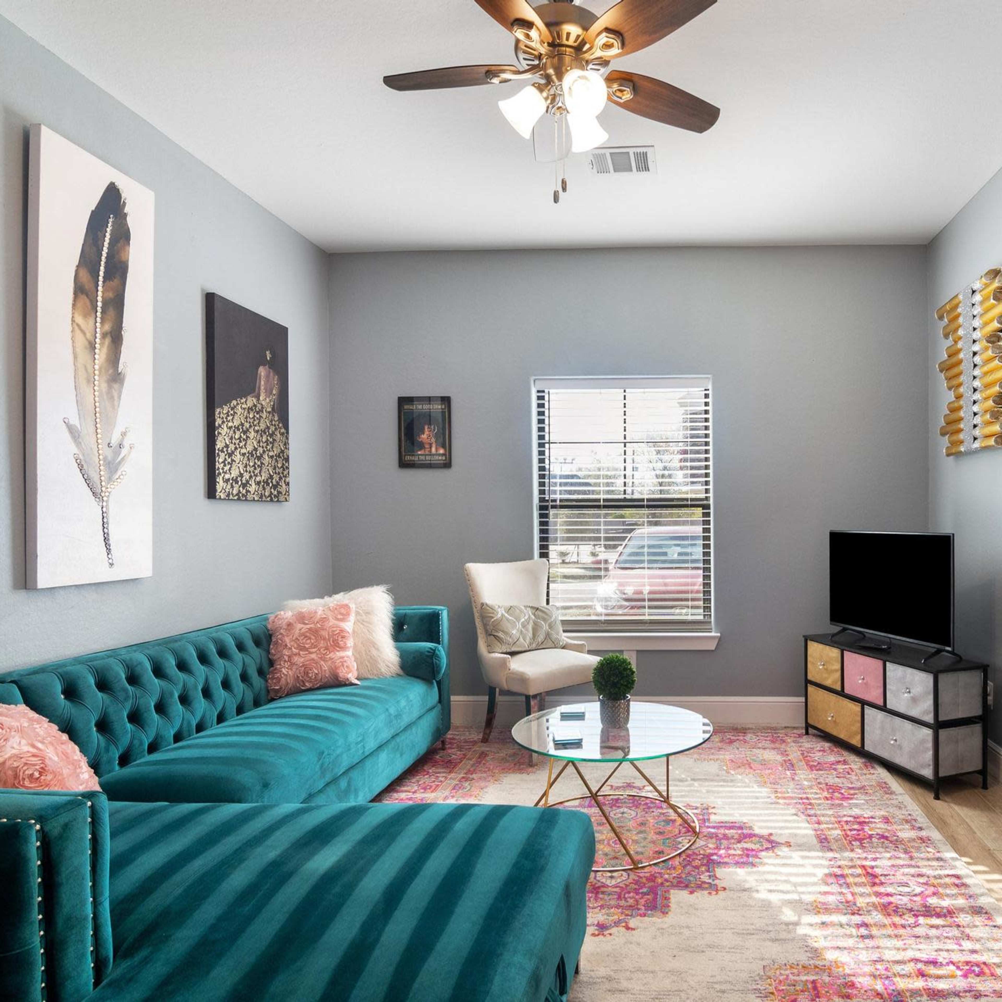 The living room features a teal velvet sofa, a round glass coffee table, and a television on a multi-drawer stand, with artwork on the walls and a window allowing natural light.