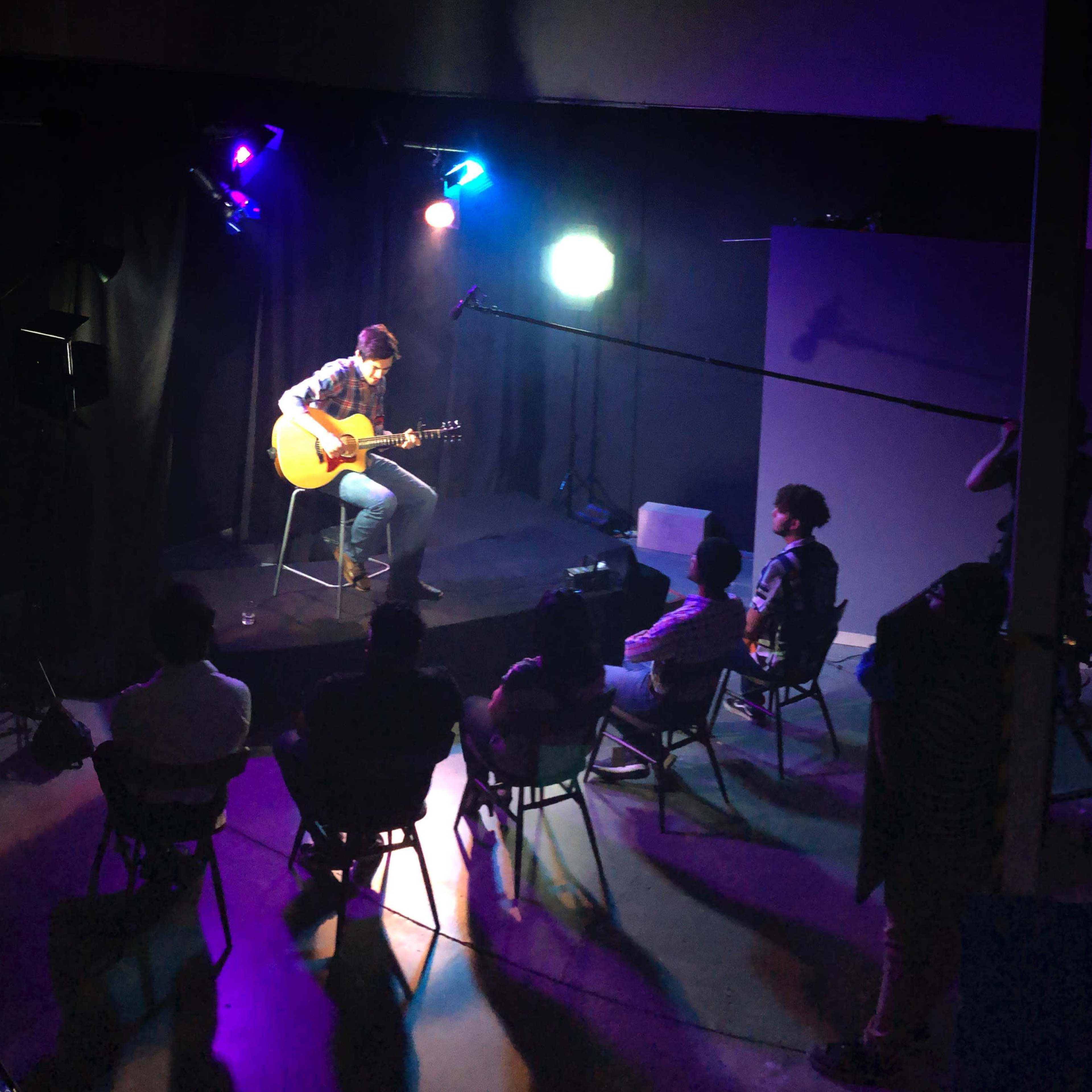 A musician plays guitar on a small stage in a dimly lit room, while an audience sits in chairs facing him.