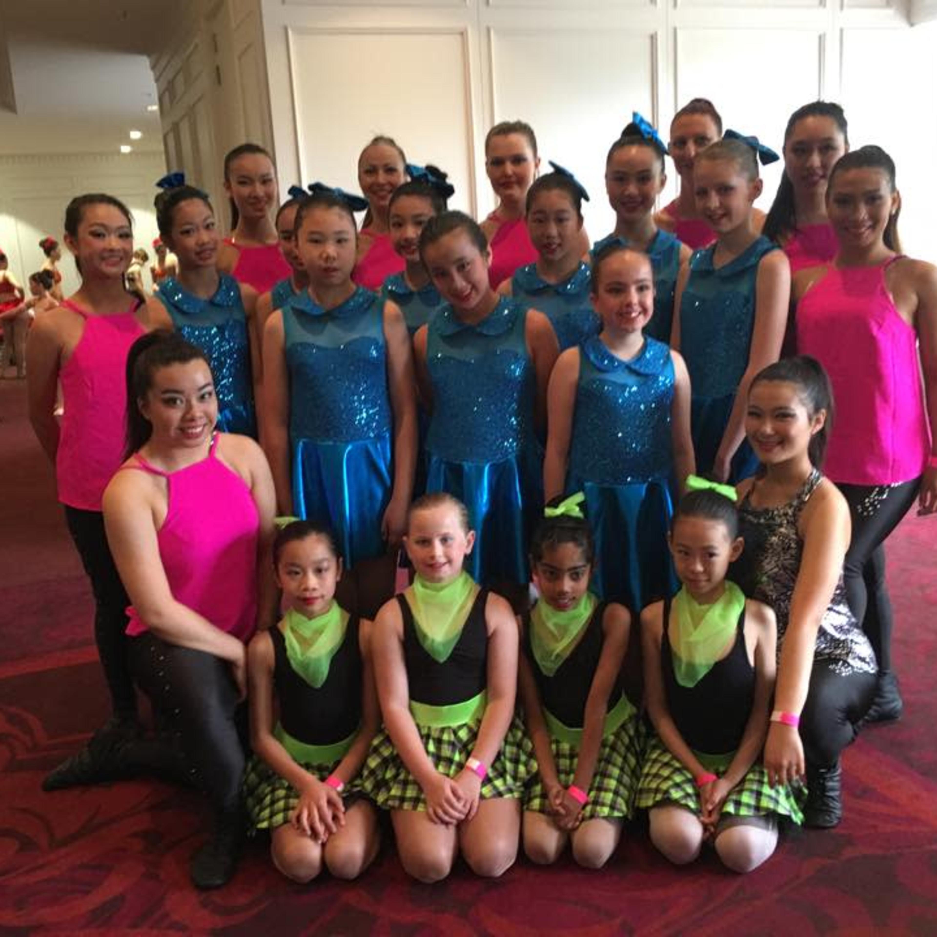 A group of young dancers in colorful costumes poses together in a well-lit interior space.