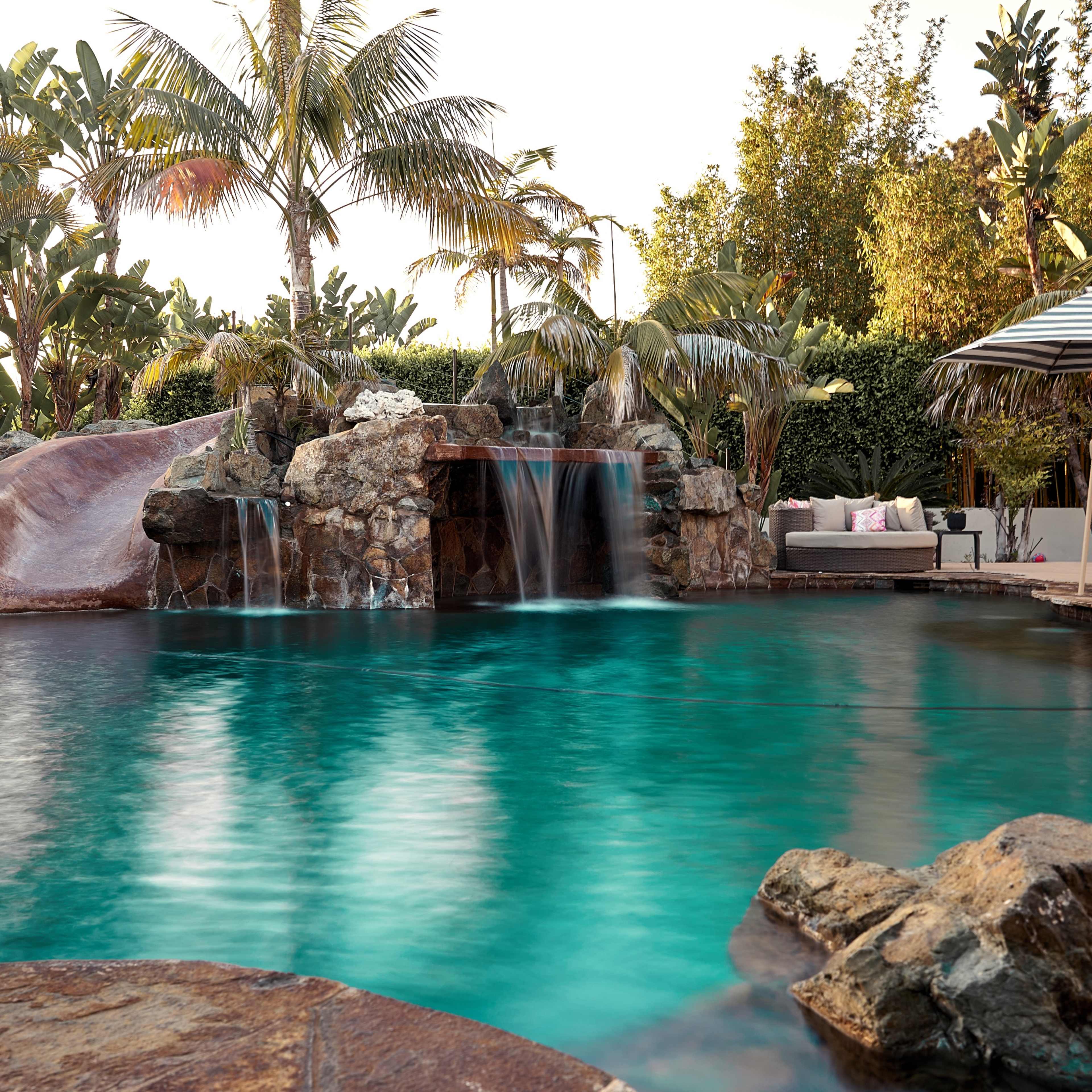 A tranquil swimming pool with cascading waterfalls, surrounded by lush tropical plants and a shaded seating area.