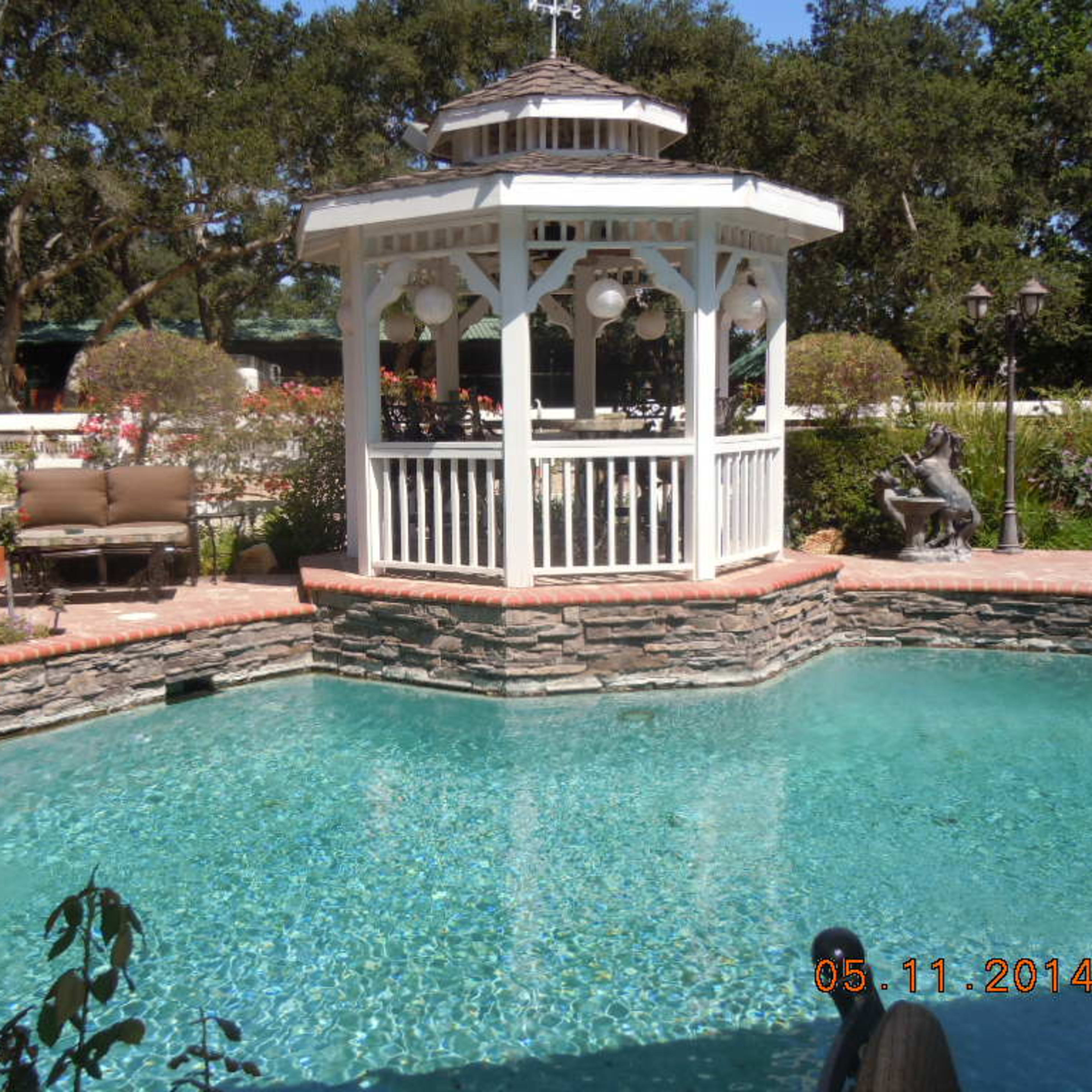 A landscaped pool area featuring a stone gazebo, surrounded by plants and seating.